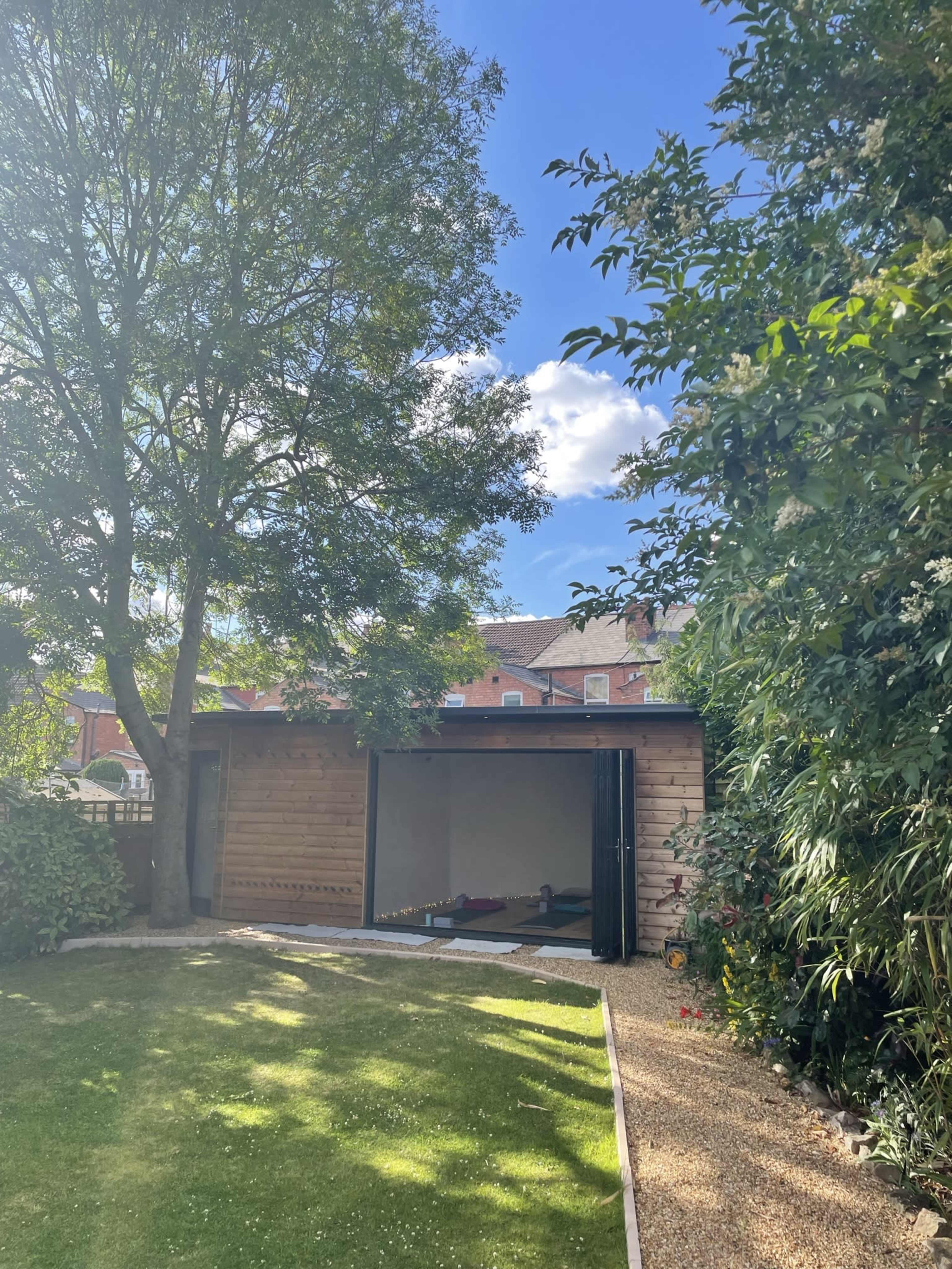 A wooden structure with sliding doors is situated in a garden surrounded by trees and shrubs under a partly cloudy sky.