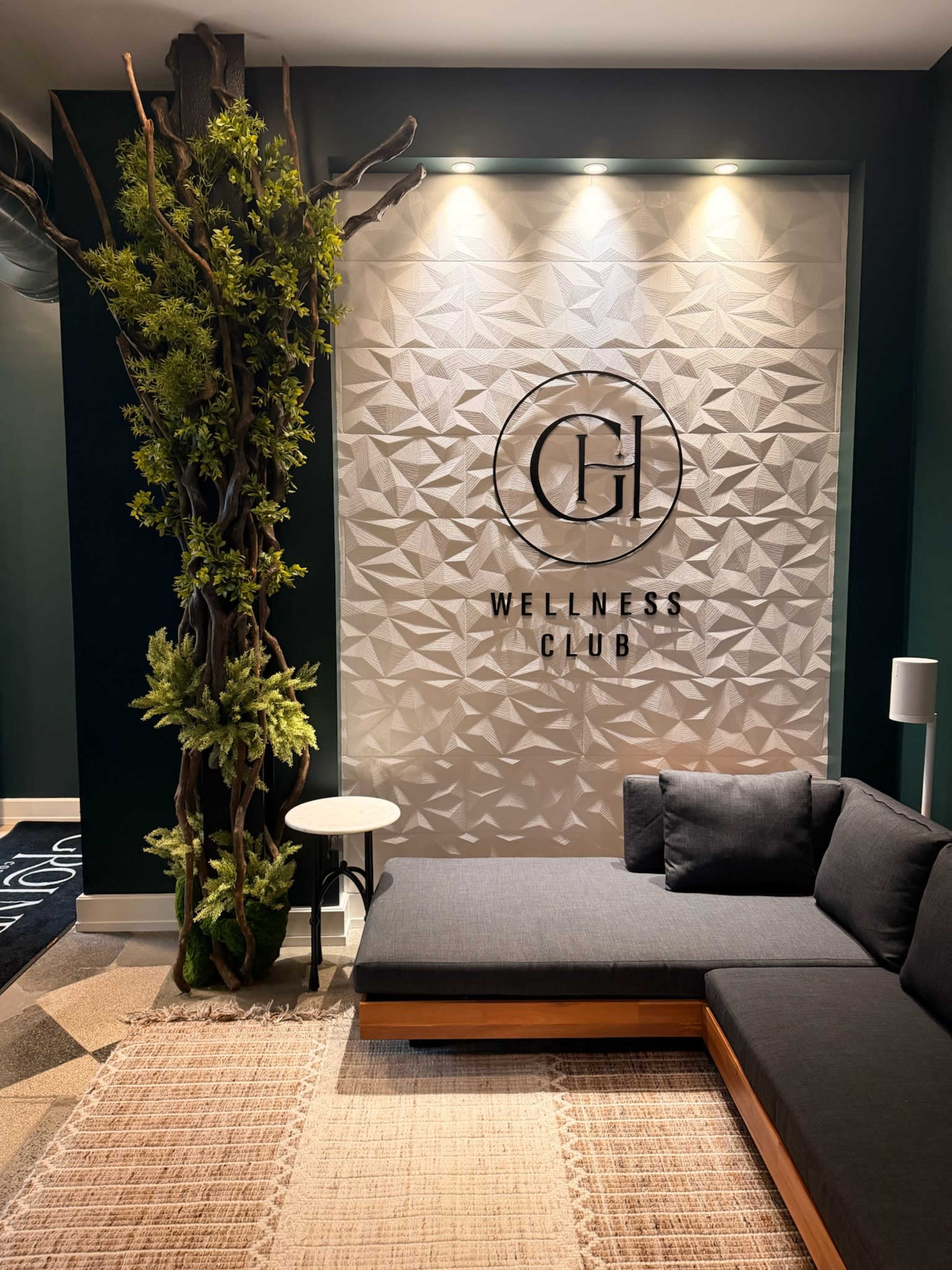 The image shows a modern wellness club entrance with a textured wall displaying the club's logo, a sofa with dark cushions, and a decorative plant.