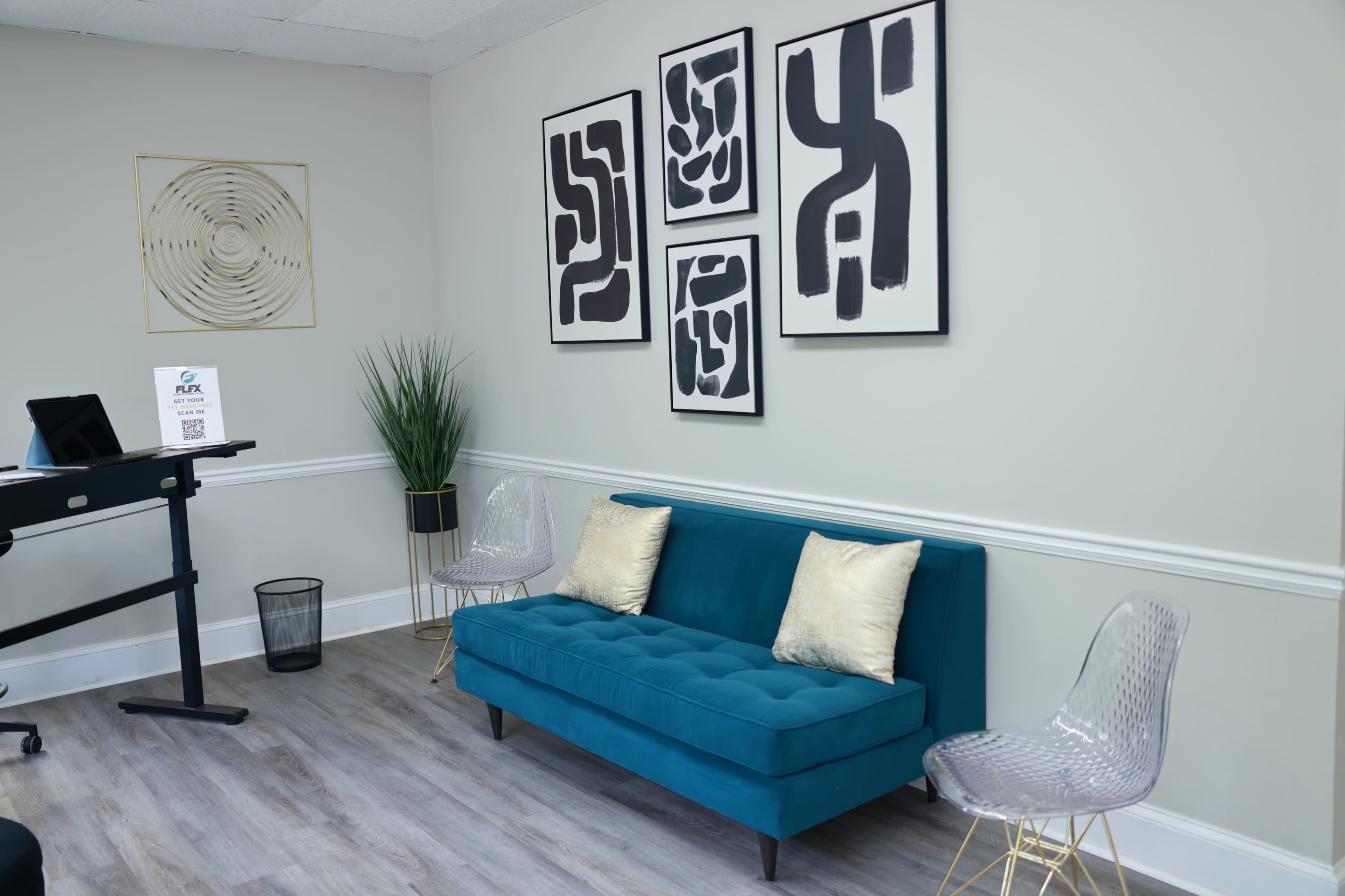 A modern waiting area featuring a teal sofa with decorative pillows, two clear acrylic chairs, and abstract wall art.