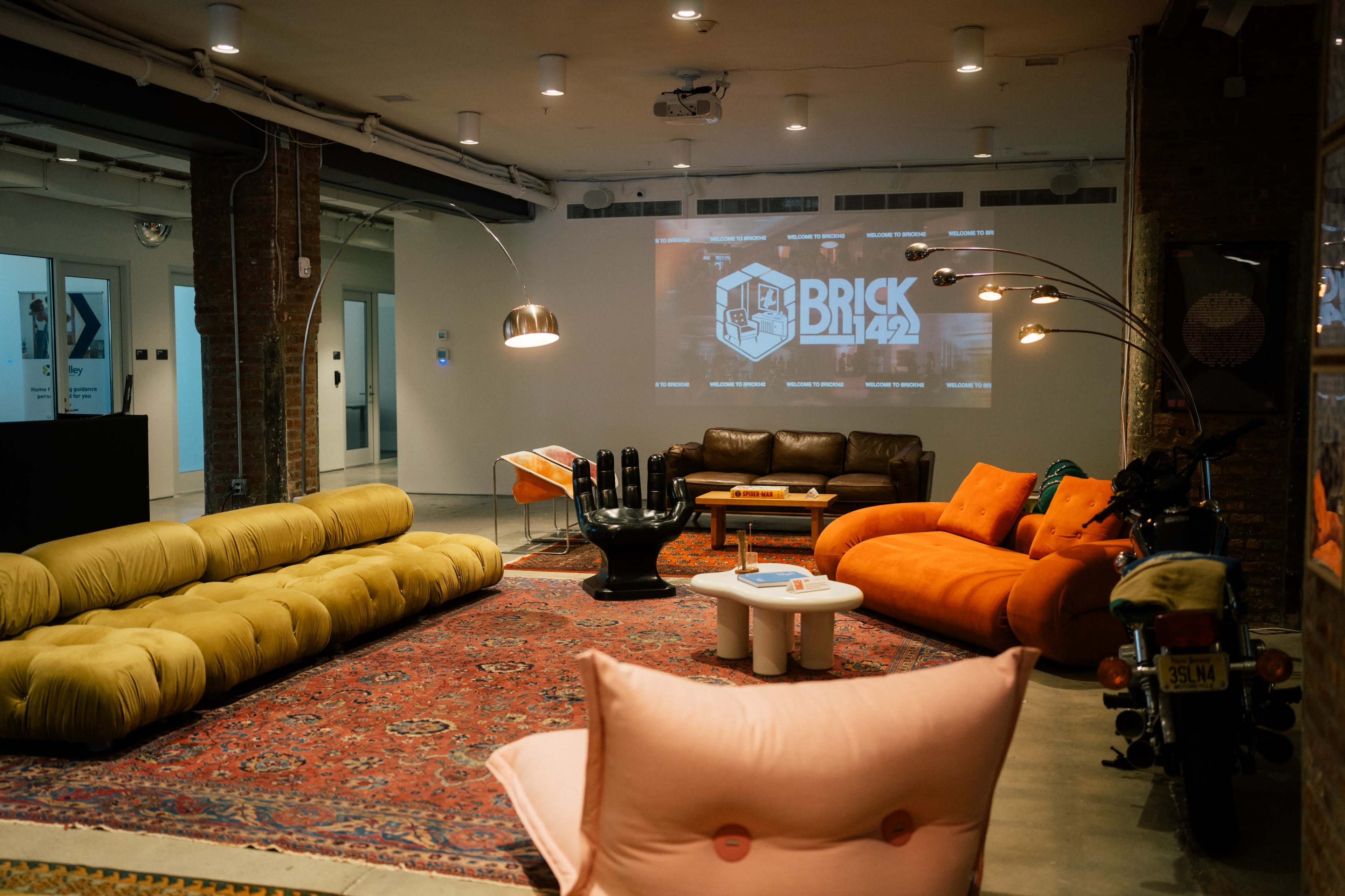 The image shows a modern lounge area featuring colorful sofas, a large area rug, a wall-mounted projector displaying "BRICK 142," and a motorcycle in the corner.