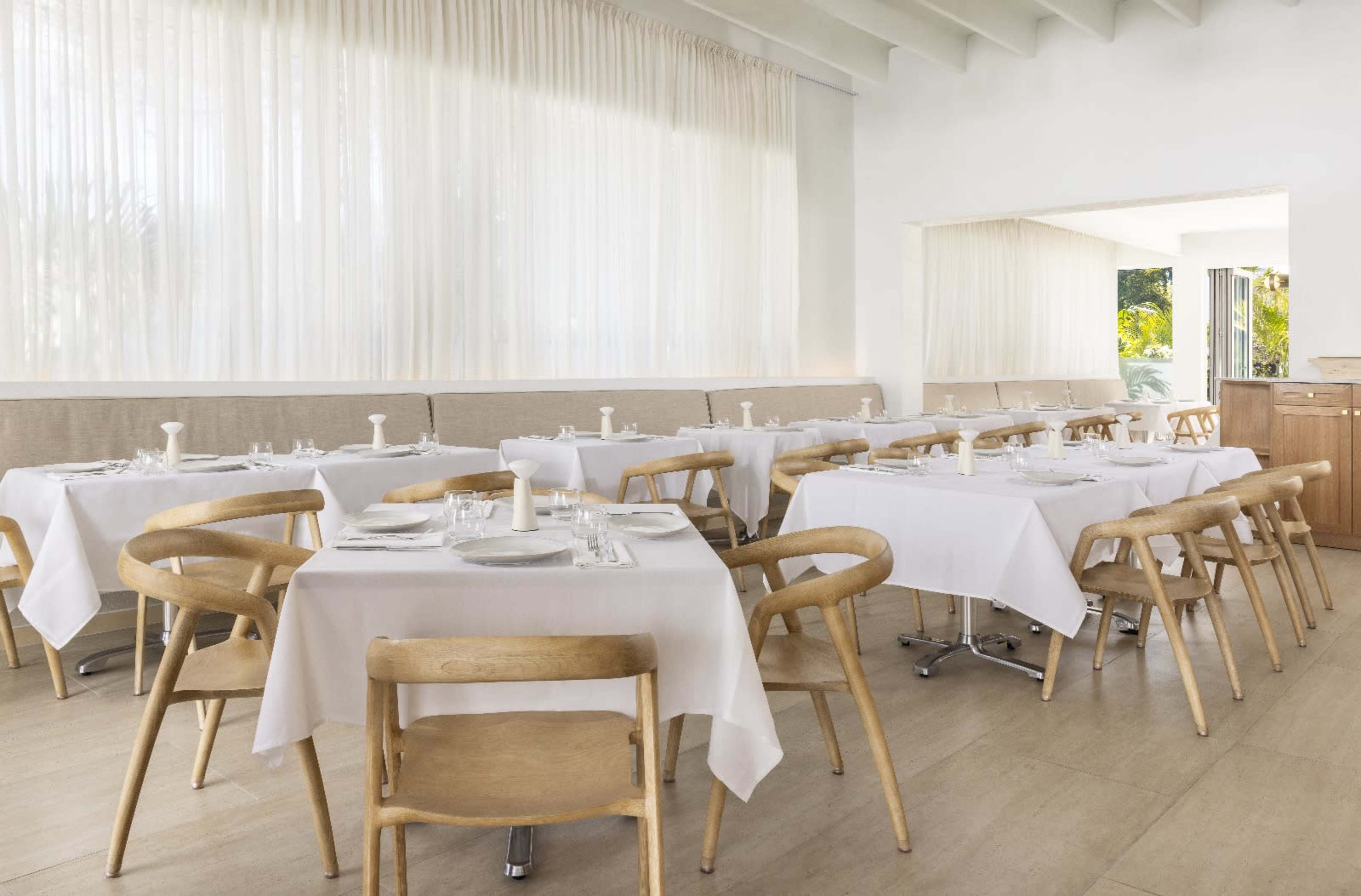 The image shows a modern restaurant interior with neatly arranged tables covered in white tablecloths, wooden chairs, and large windows allowing natural light to fill the space.