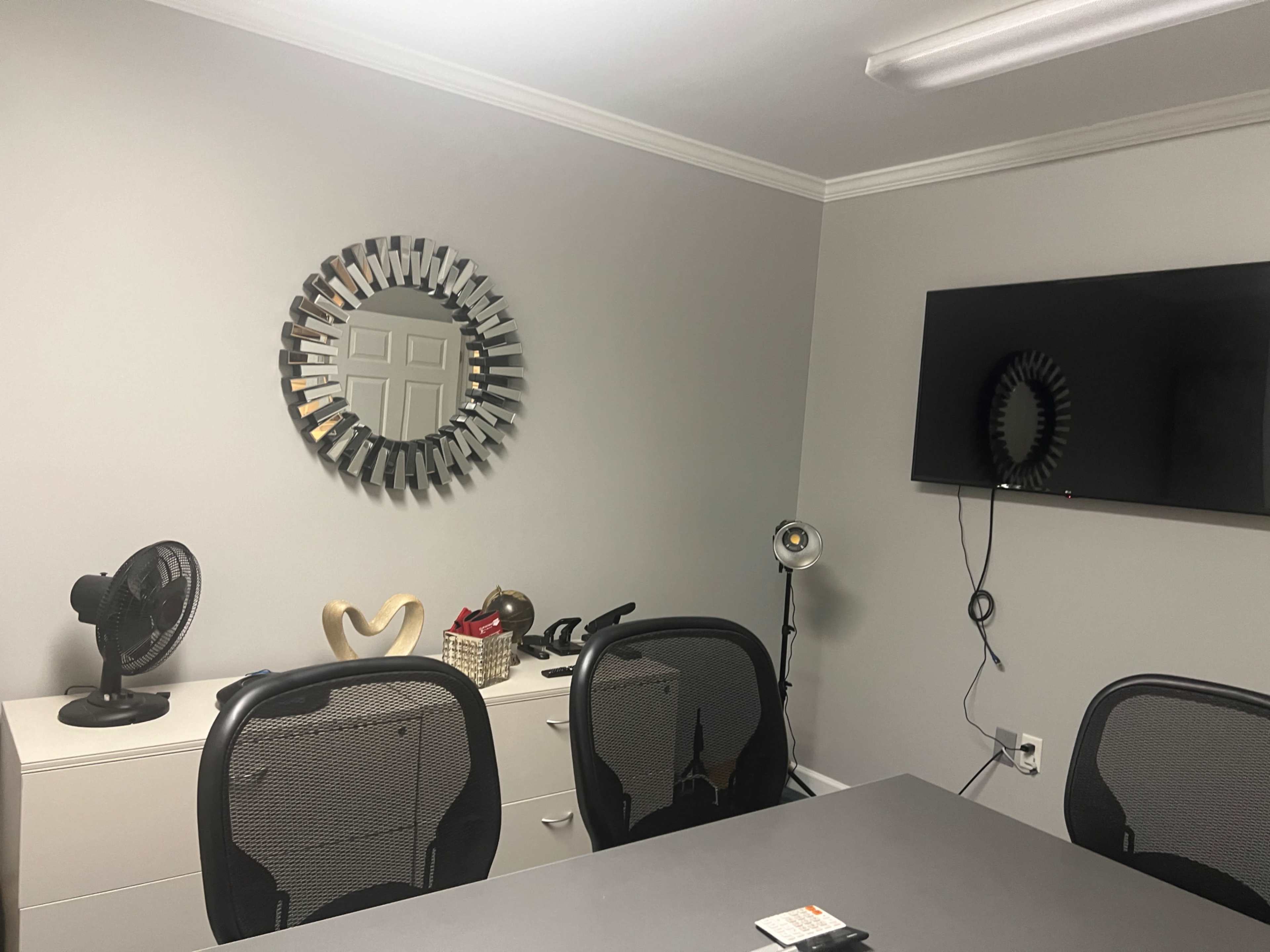 The image shows a modern office setting with a round mirror on the wall, a television, a fan, and a grey table surrounded by black mesh chairs.