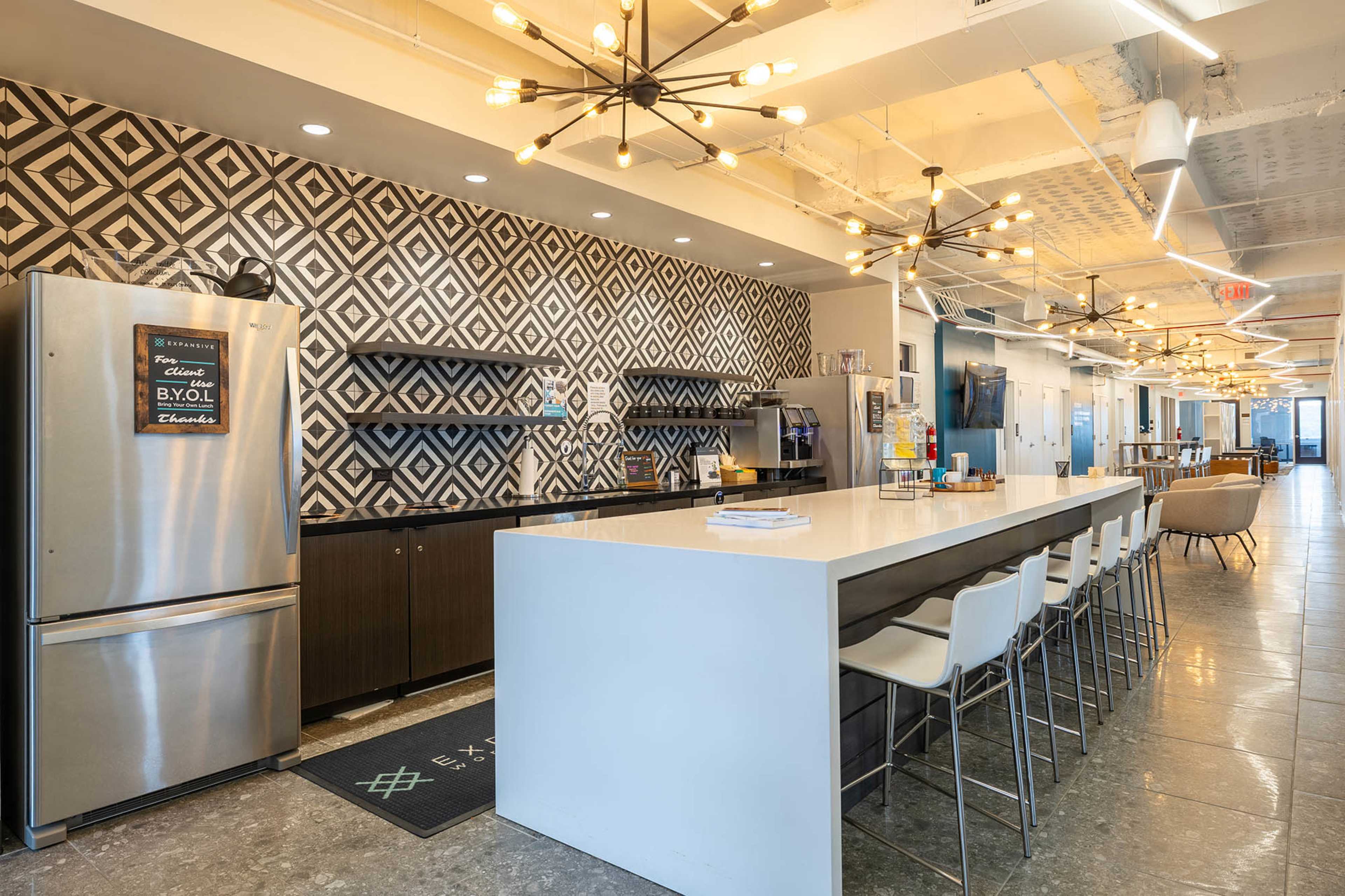 The image shows a modern kitchen and lounge area with a large white countertop, bar seating, and a patterned wall behind the kitchen appliances.