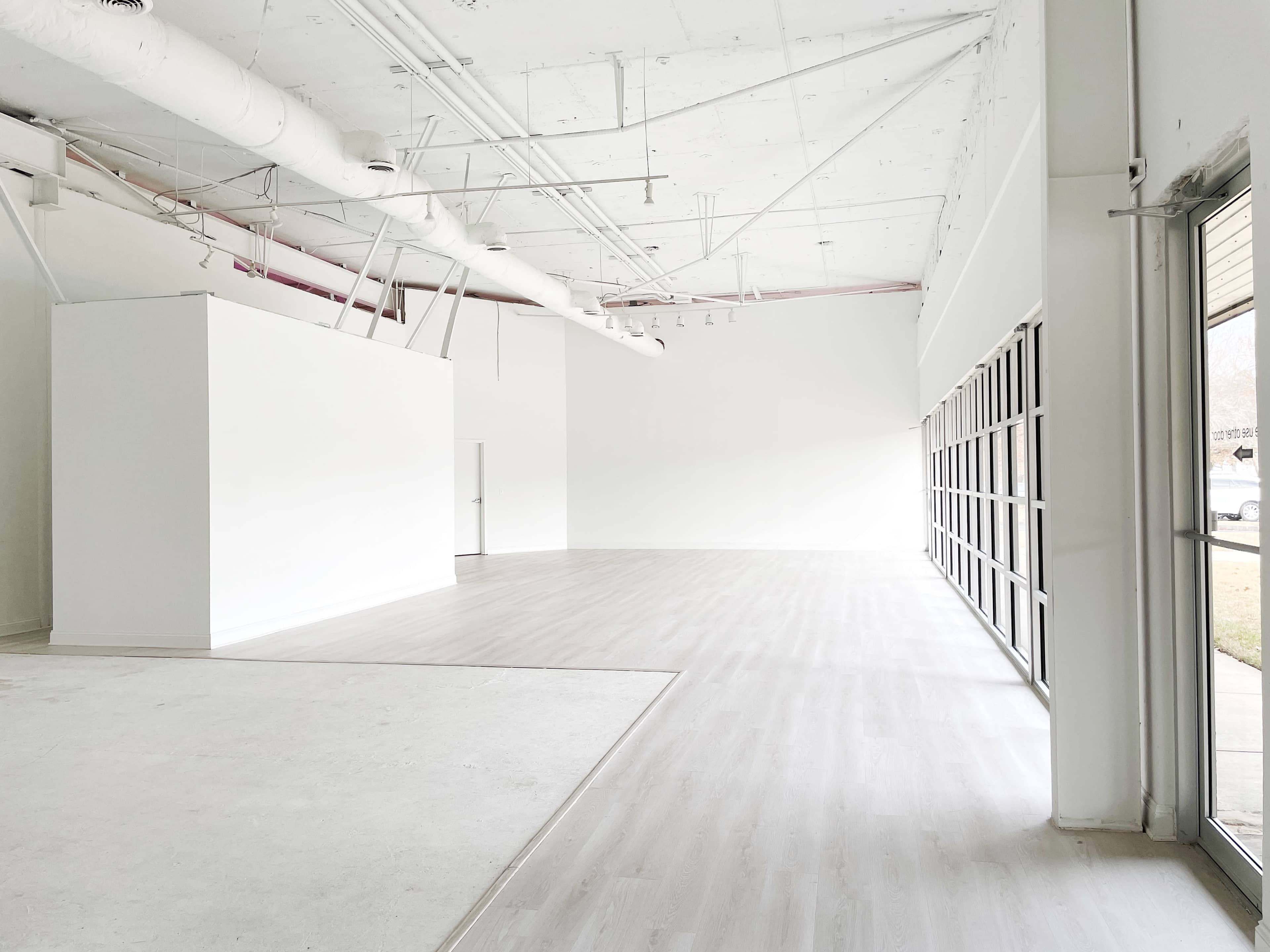 A spacious, empty room with white walls, large windows, and light-colored flooring.