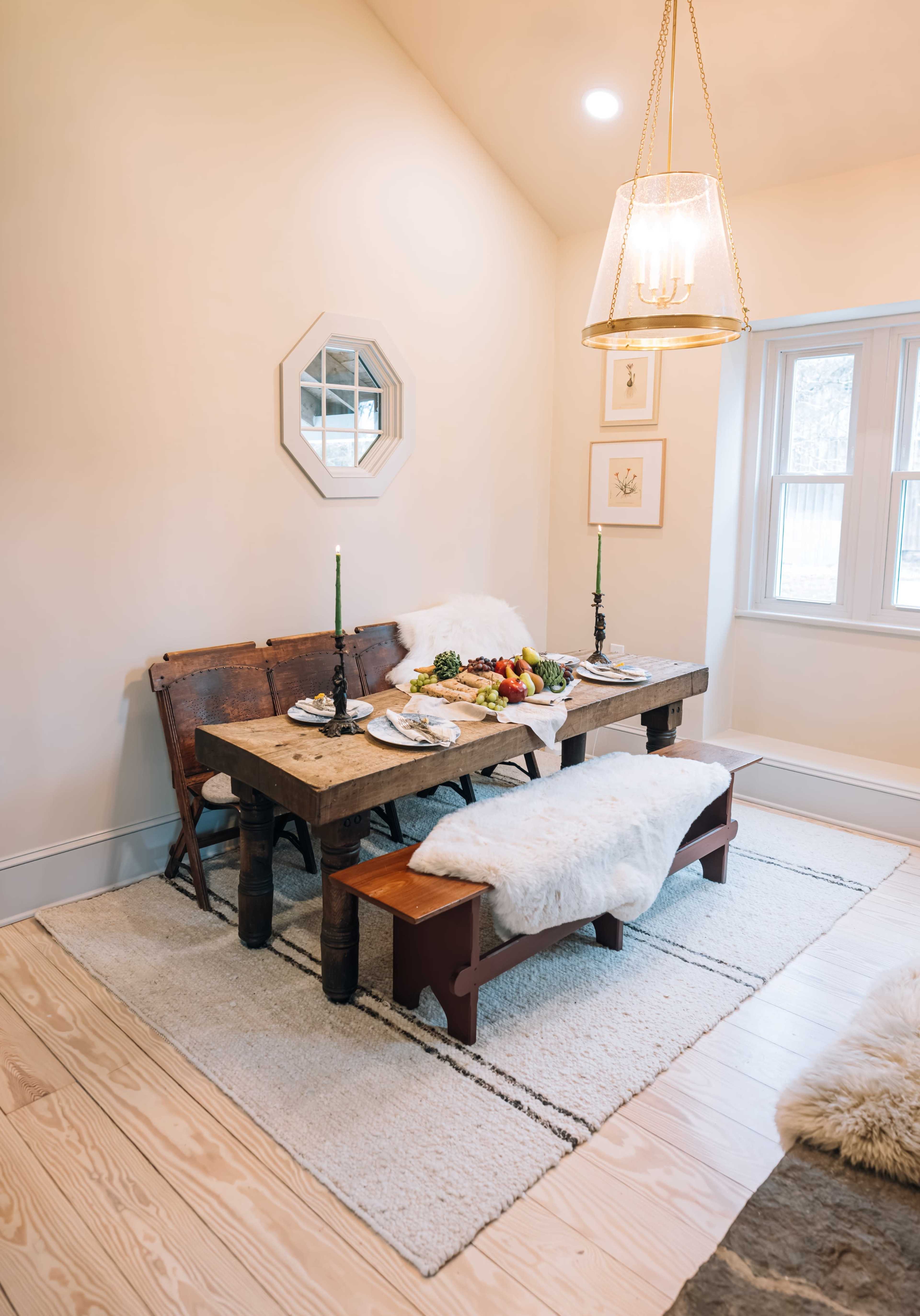 A rustic dining table set with dishes and a centerpiece is surrounded by benches covered in soft textiles in a well-lit room.