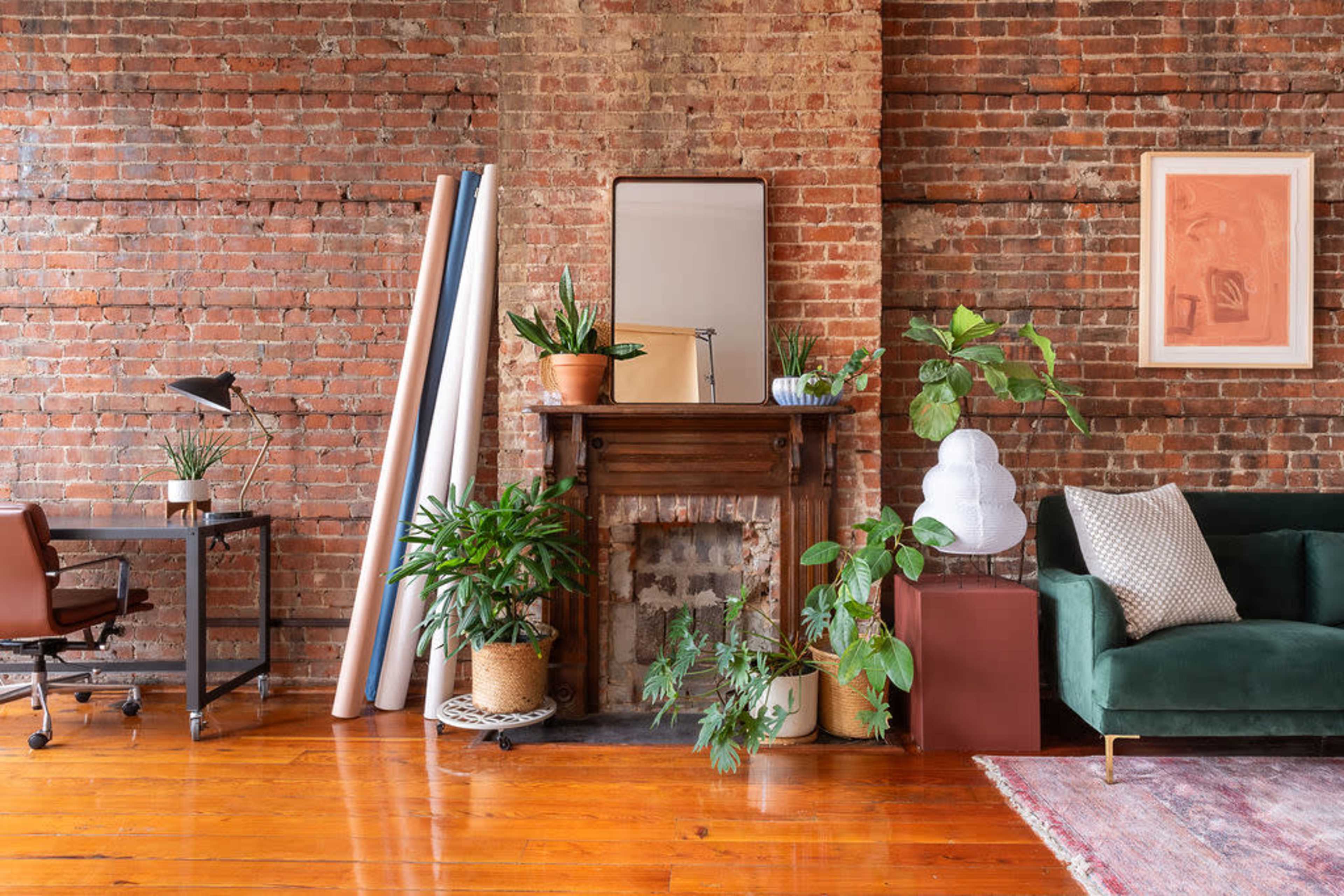 A cozy interior space featuring a brick wall, a green sofa, a desk with an office chair, plants, and a fireplace with decorative items.