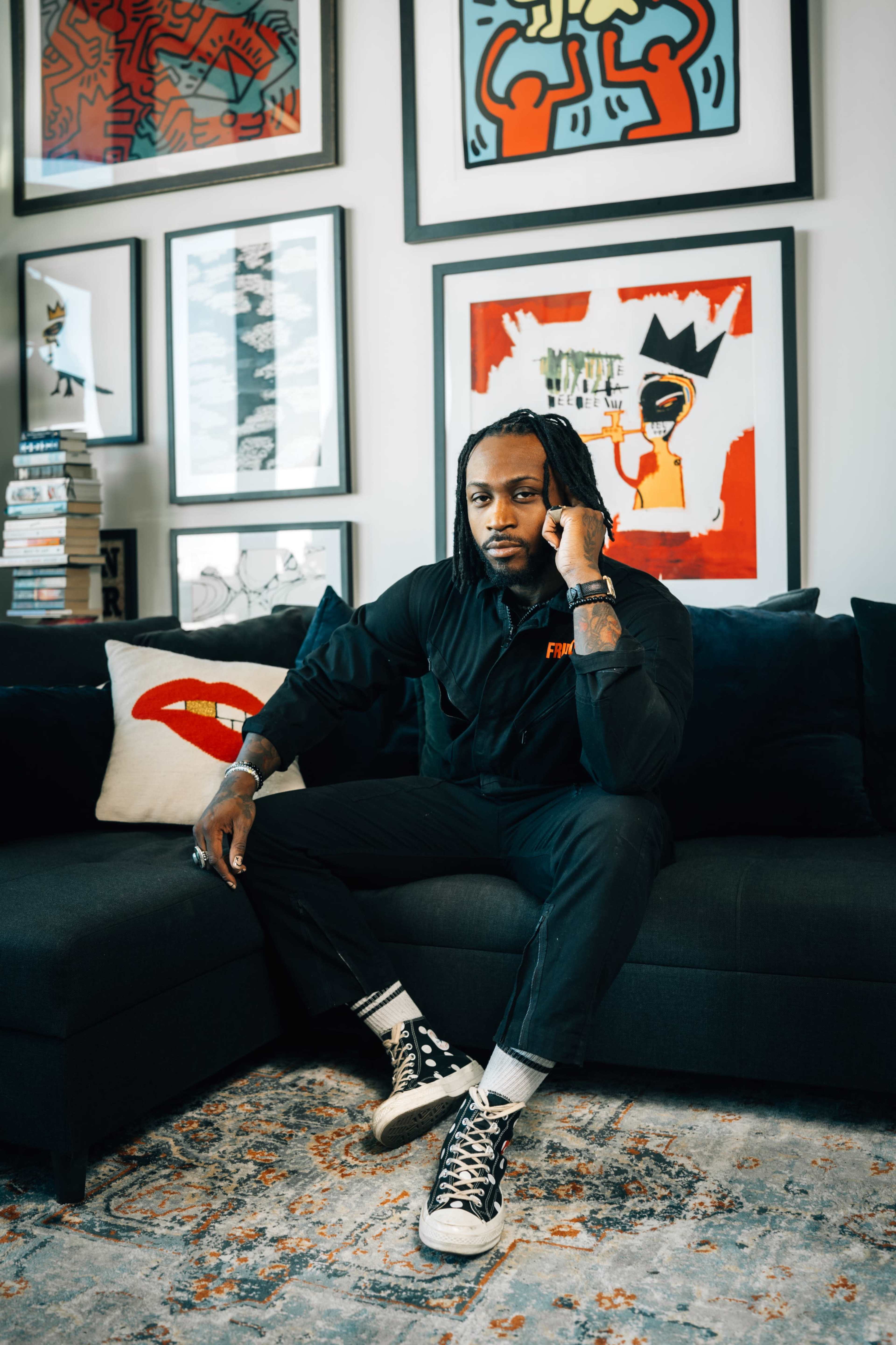 A man with long hair sits on a dark couch in a stylish room adorned with colorful artwork and a stack of books.
