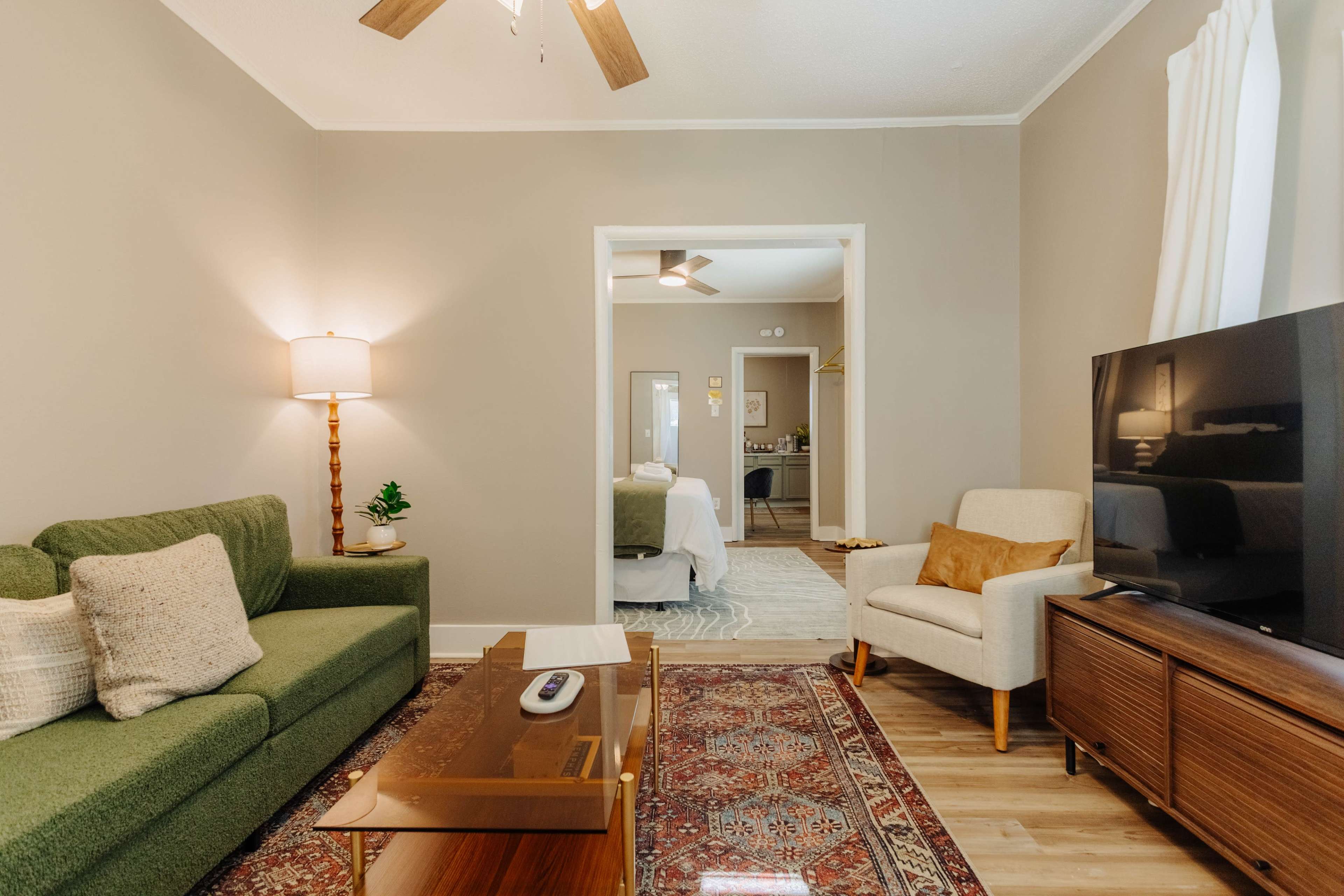 A cozy living room features a green sofa, a wooden coffee table, and a television, with a doorway leading to a light-colored bedroom in the background.