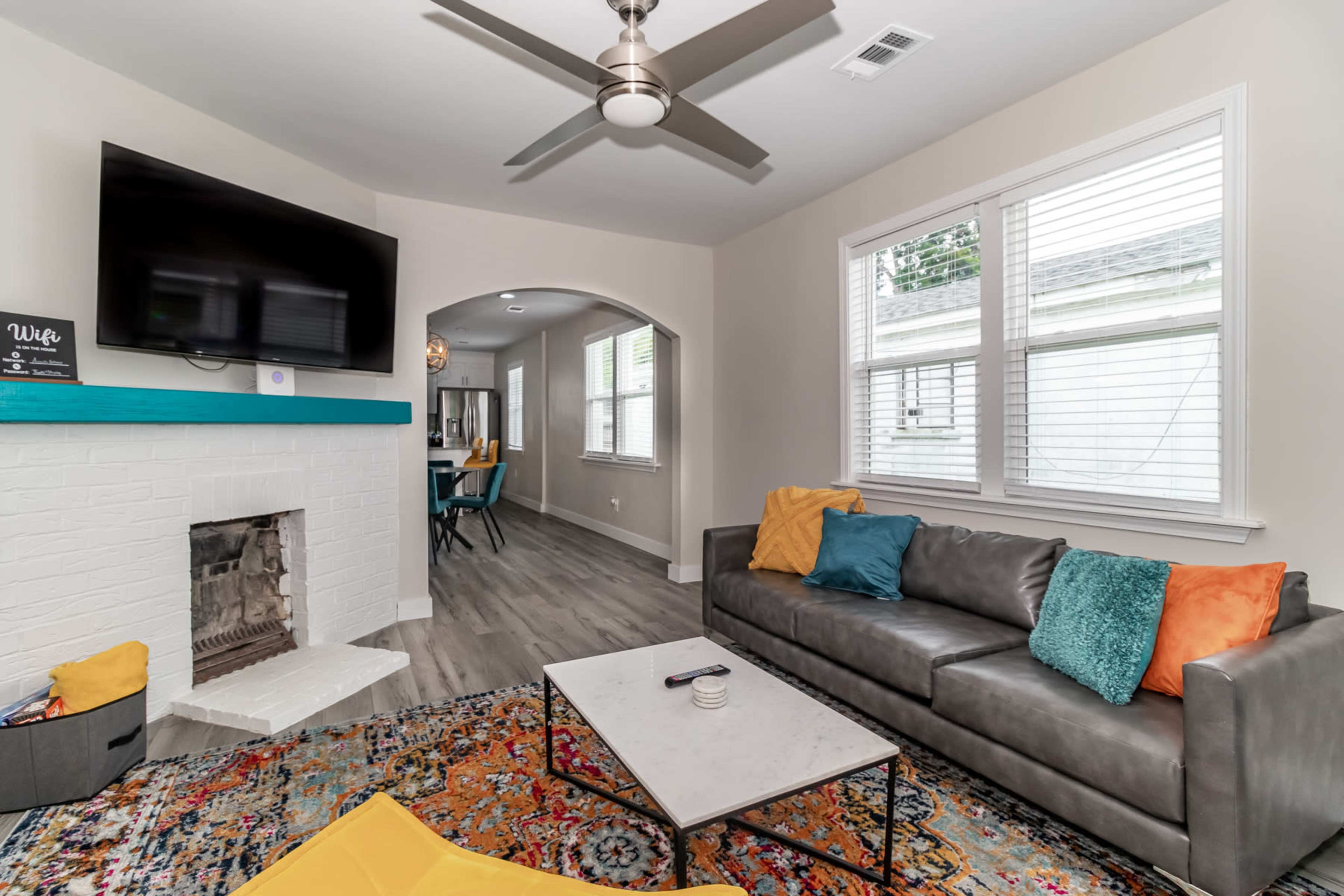 The living room features a gray sofa with colorful pillows, a coffee table on a patterned rug, and a television mounted above a white brick fireplace.
