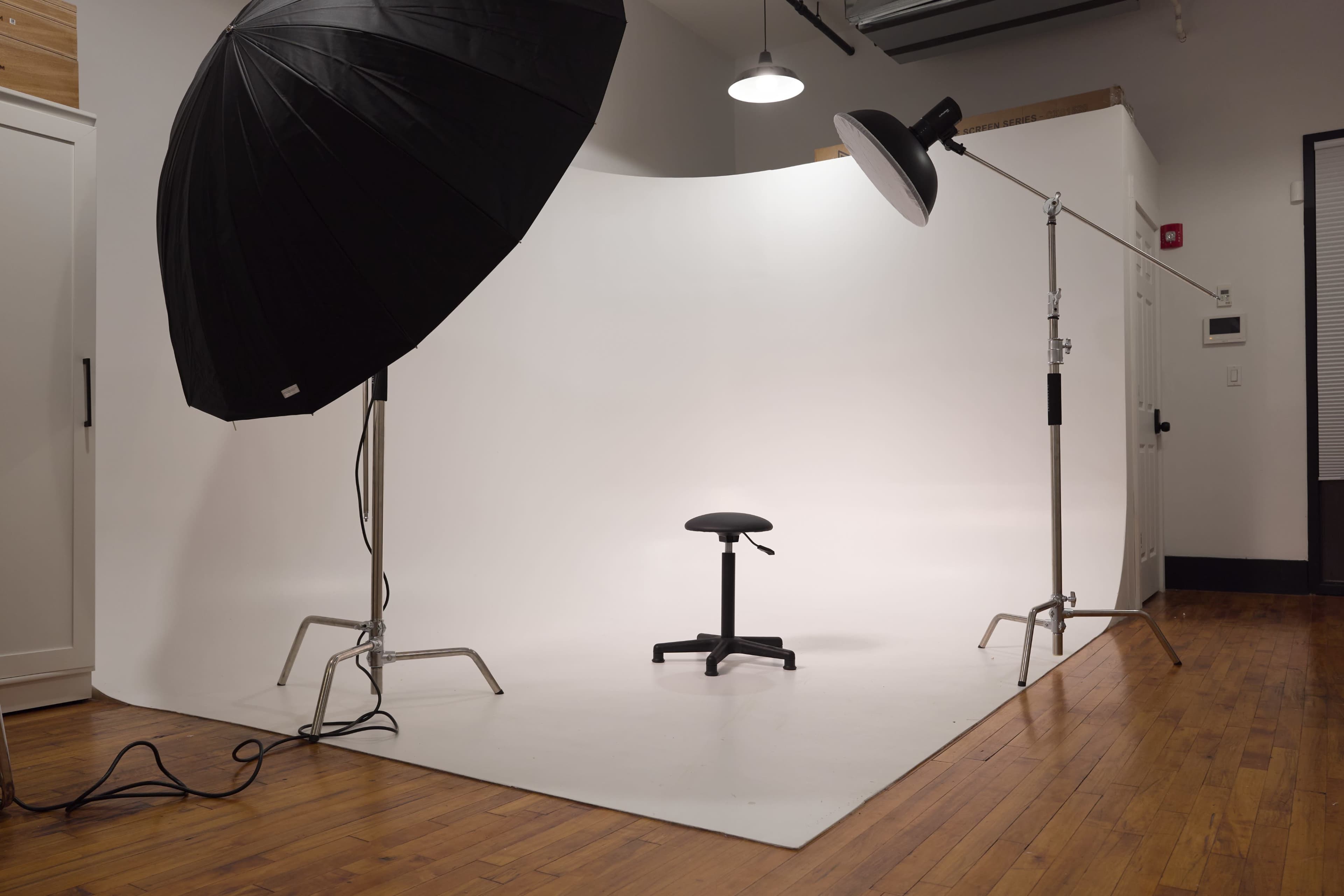 A photography studio features a large black umbrella light and a softbox positioned above a plain white backdrop with an empty stool in the center.