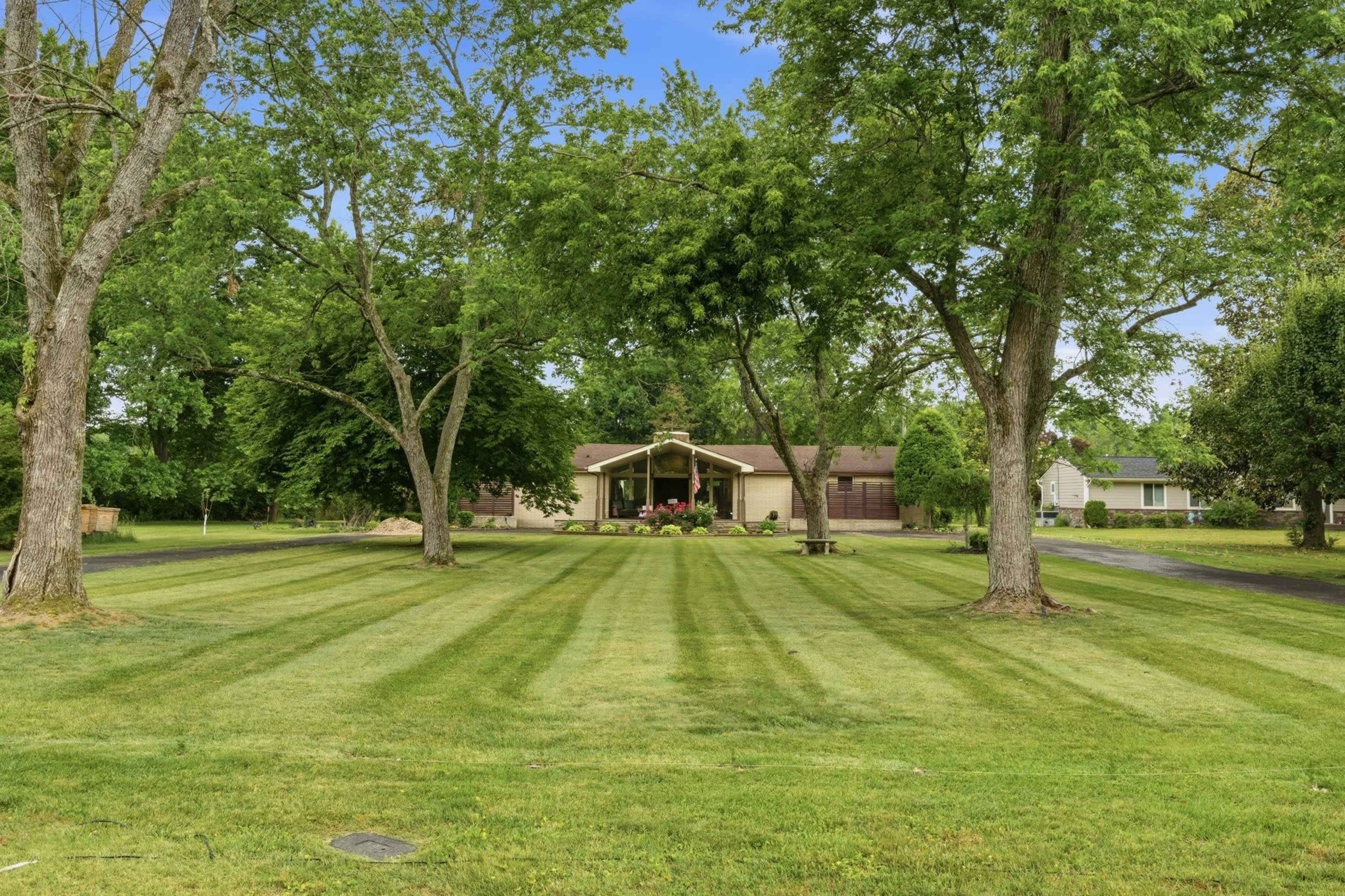 A single-story house with a well-maintained lawn and trees is set in a landscaped area featuring trimmed grass and a clear blue sky.