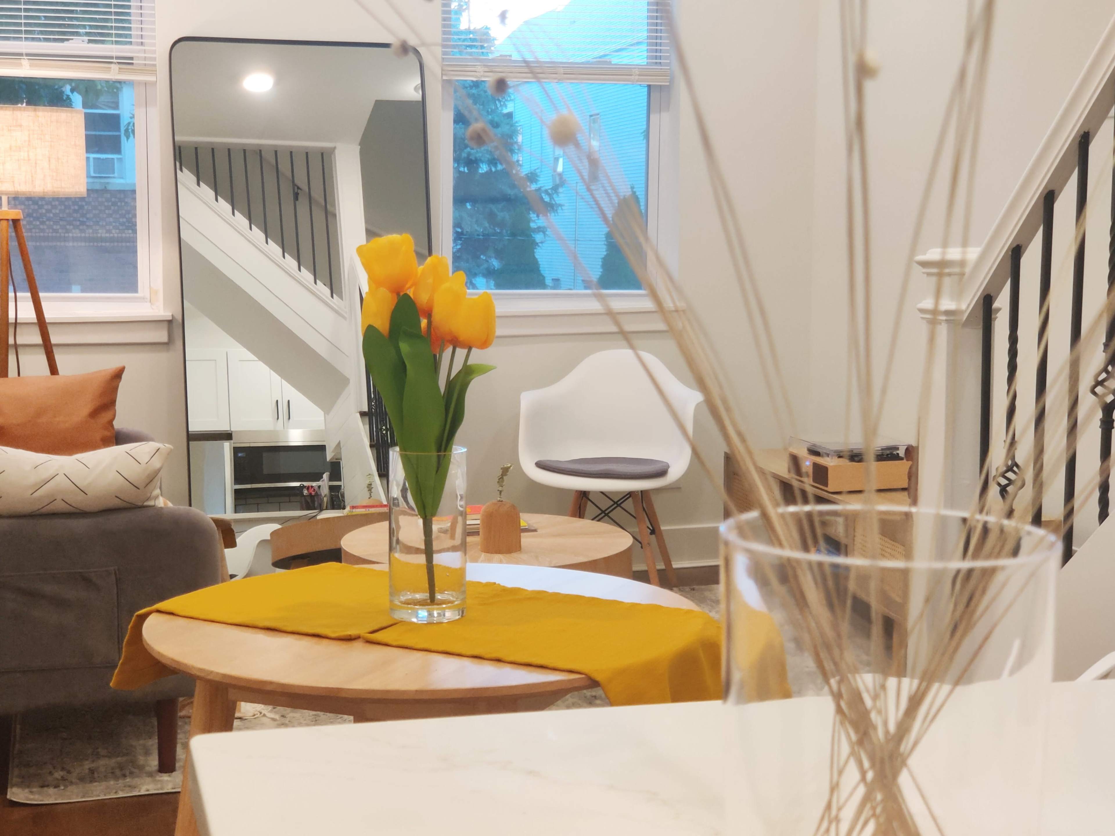 A cozy living room featuring a vase of yellow tulips on a wooden coffee table, with a staircase and kitchen visible in the background.