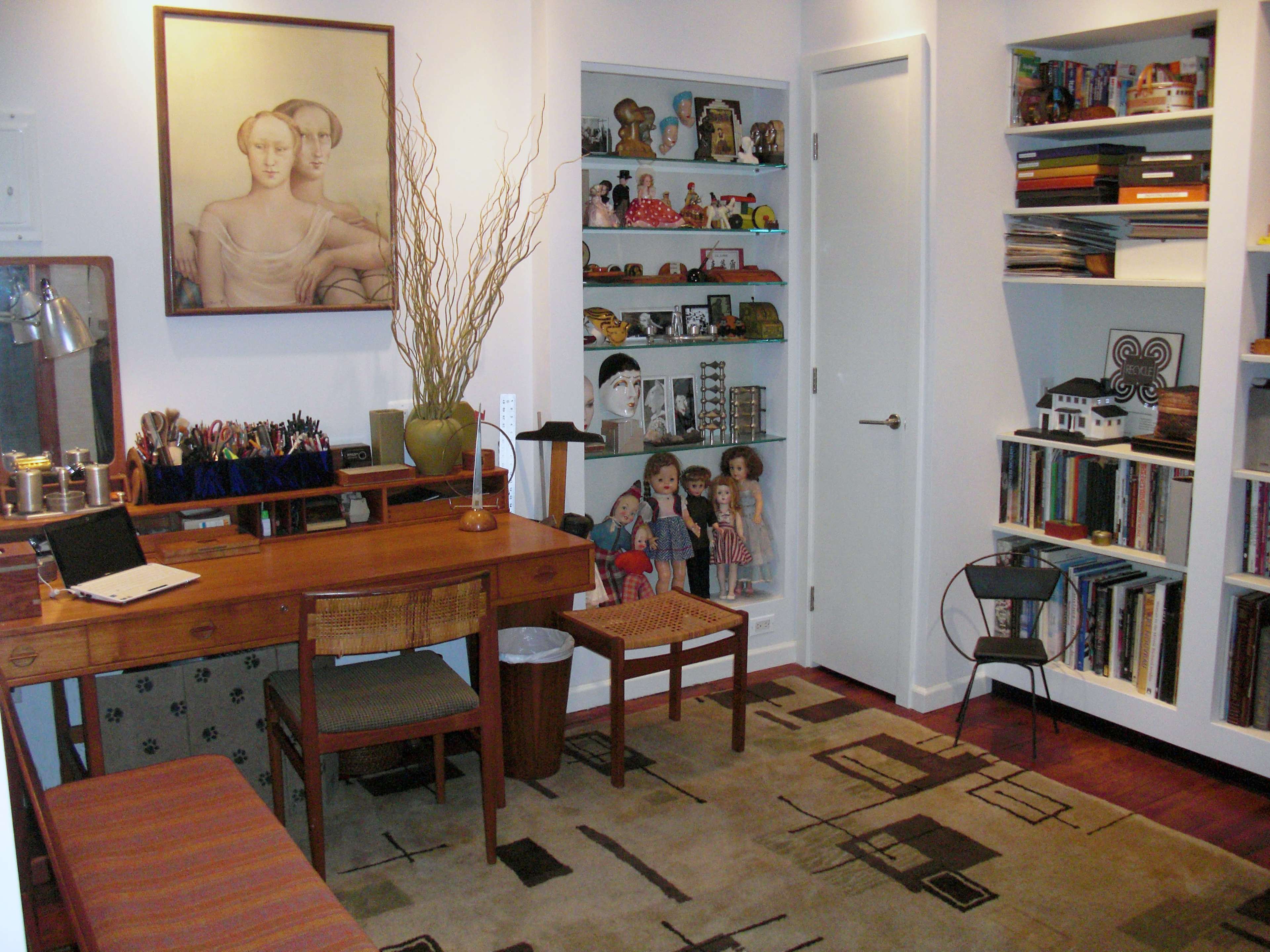 A cozy workspace featuring a wooden desk with art supplies, a seating area, shelves filled with books and decorative items, and a painting on the wall.