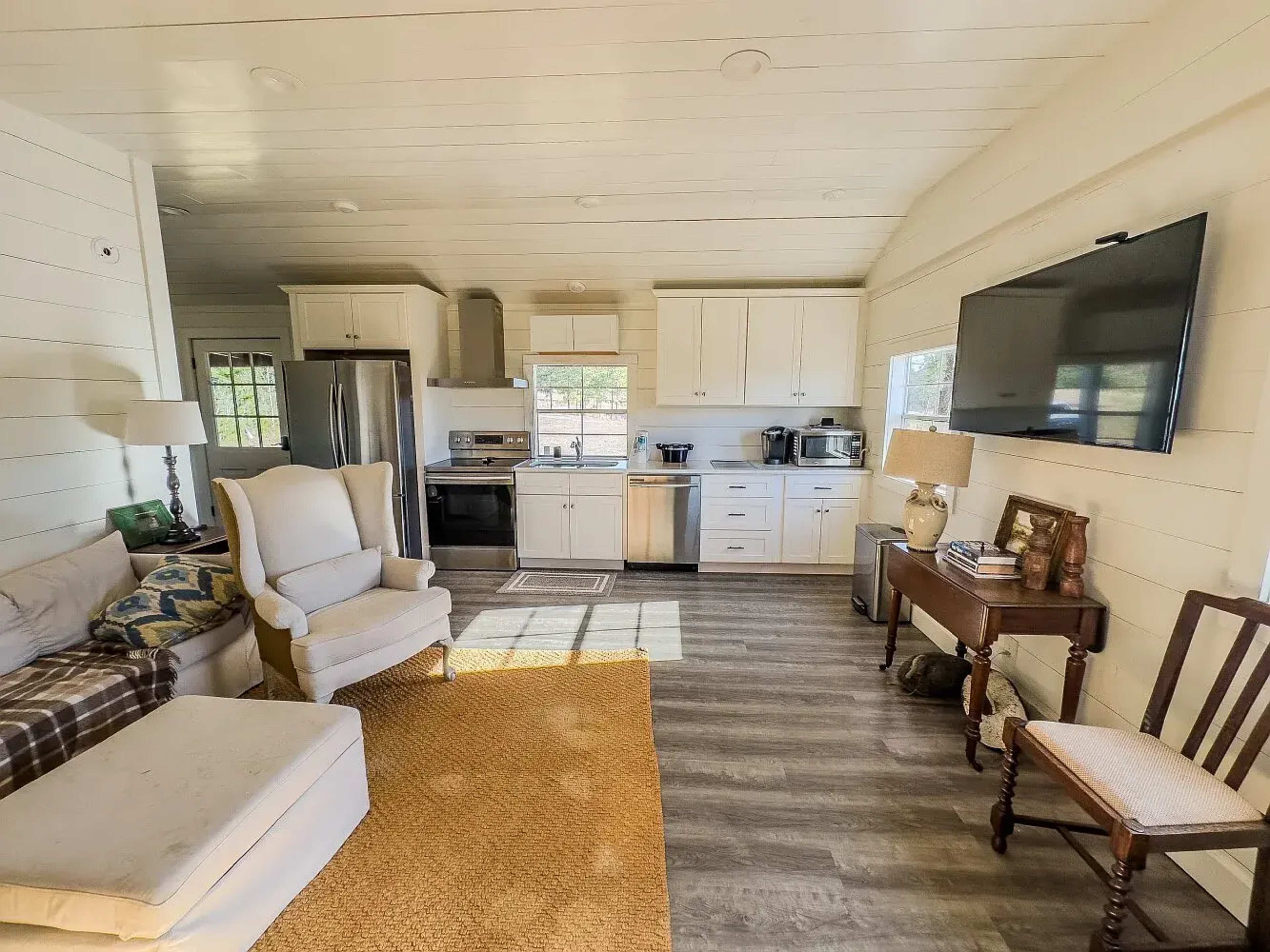 The image shows a cozy, open-concept living area featuring a light-colored sofa, a small dining table, and a kitchen with modern appliances.