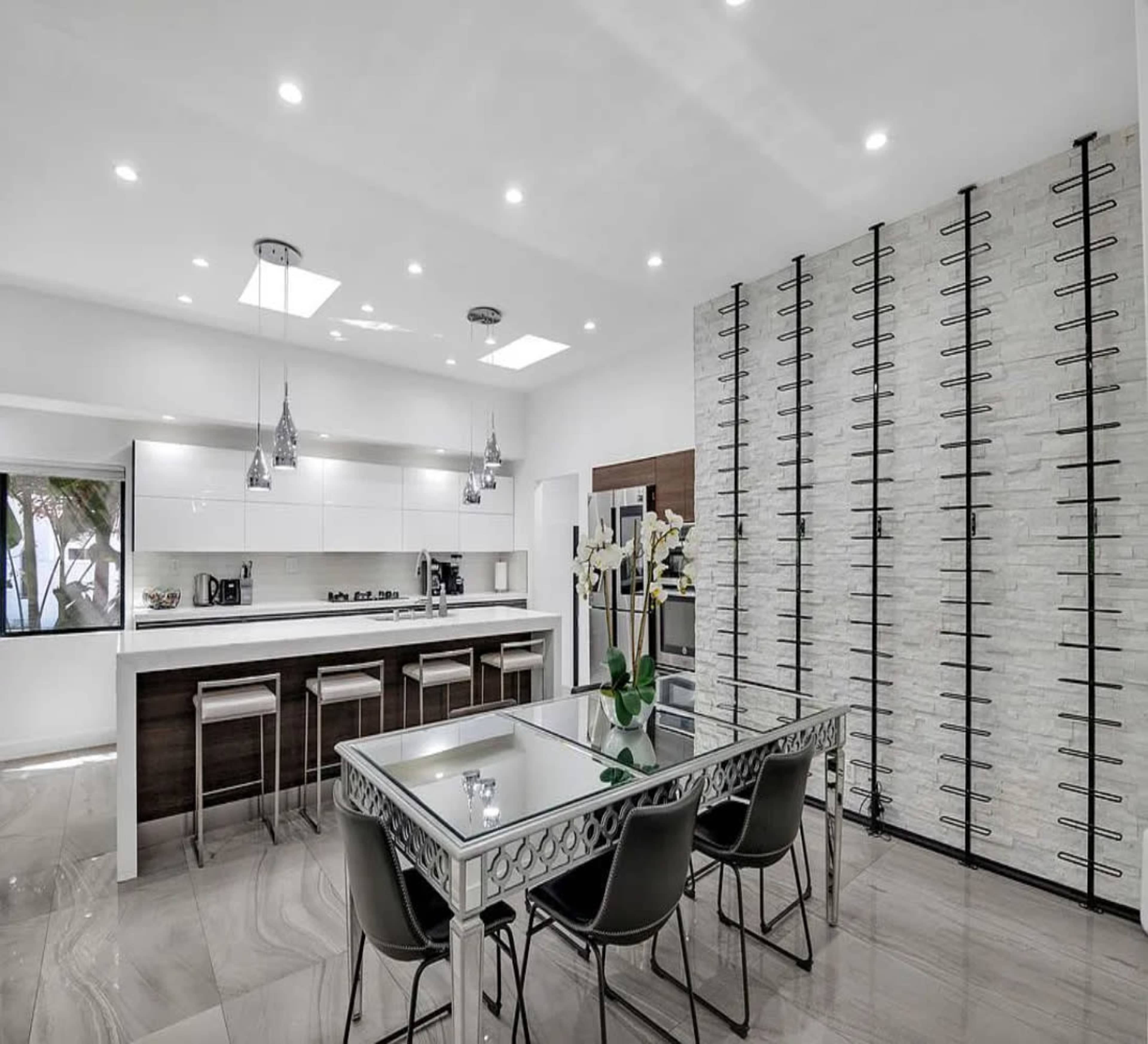 The image shows a modern kitchen and dining area featuring a sleek table, metallic chairs, and a textured wall, illuminated by recessed lighting.
