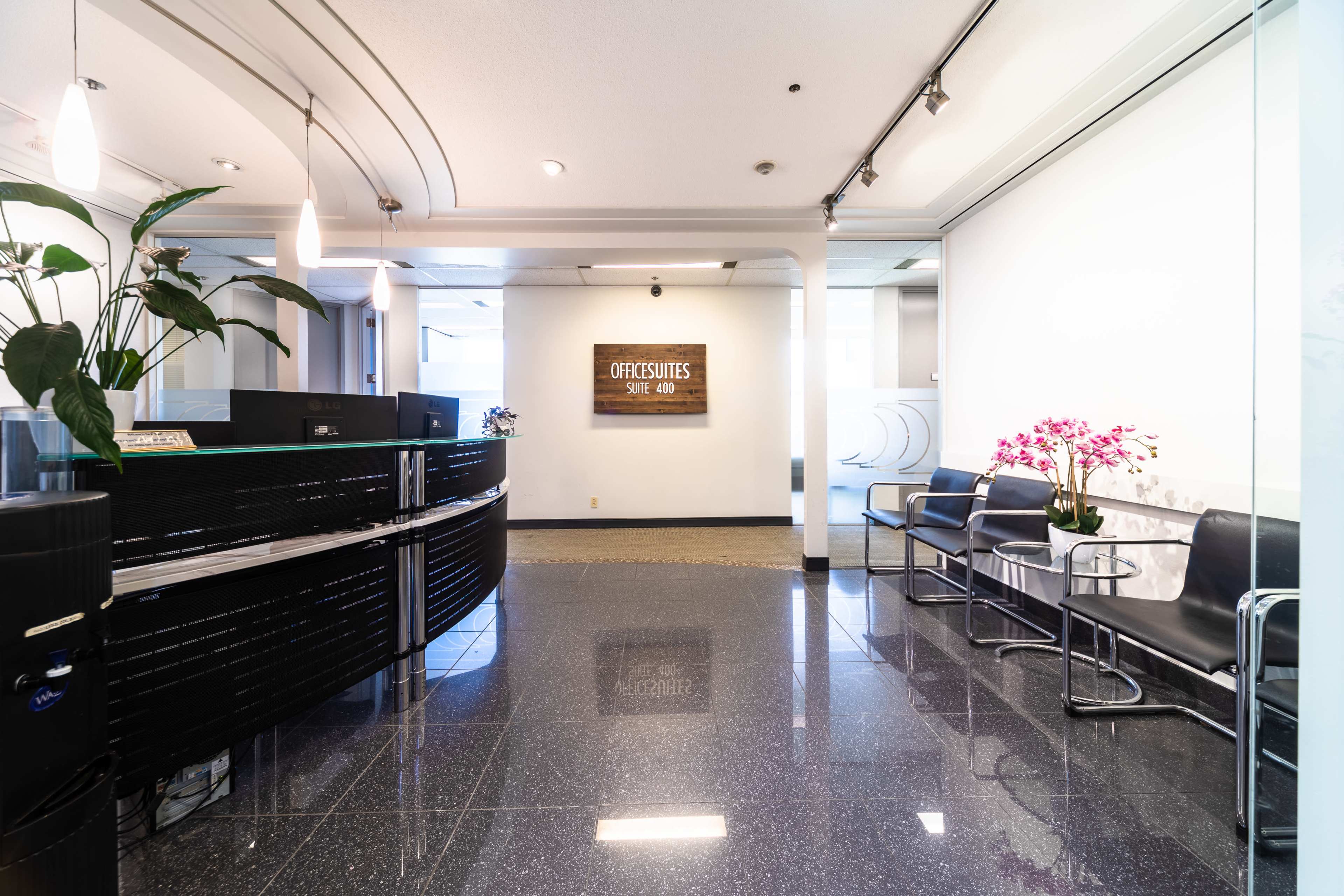 A modern office reception area with a sleek front desk, a row of black chairs, and decorative plants.