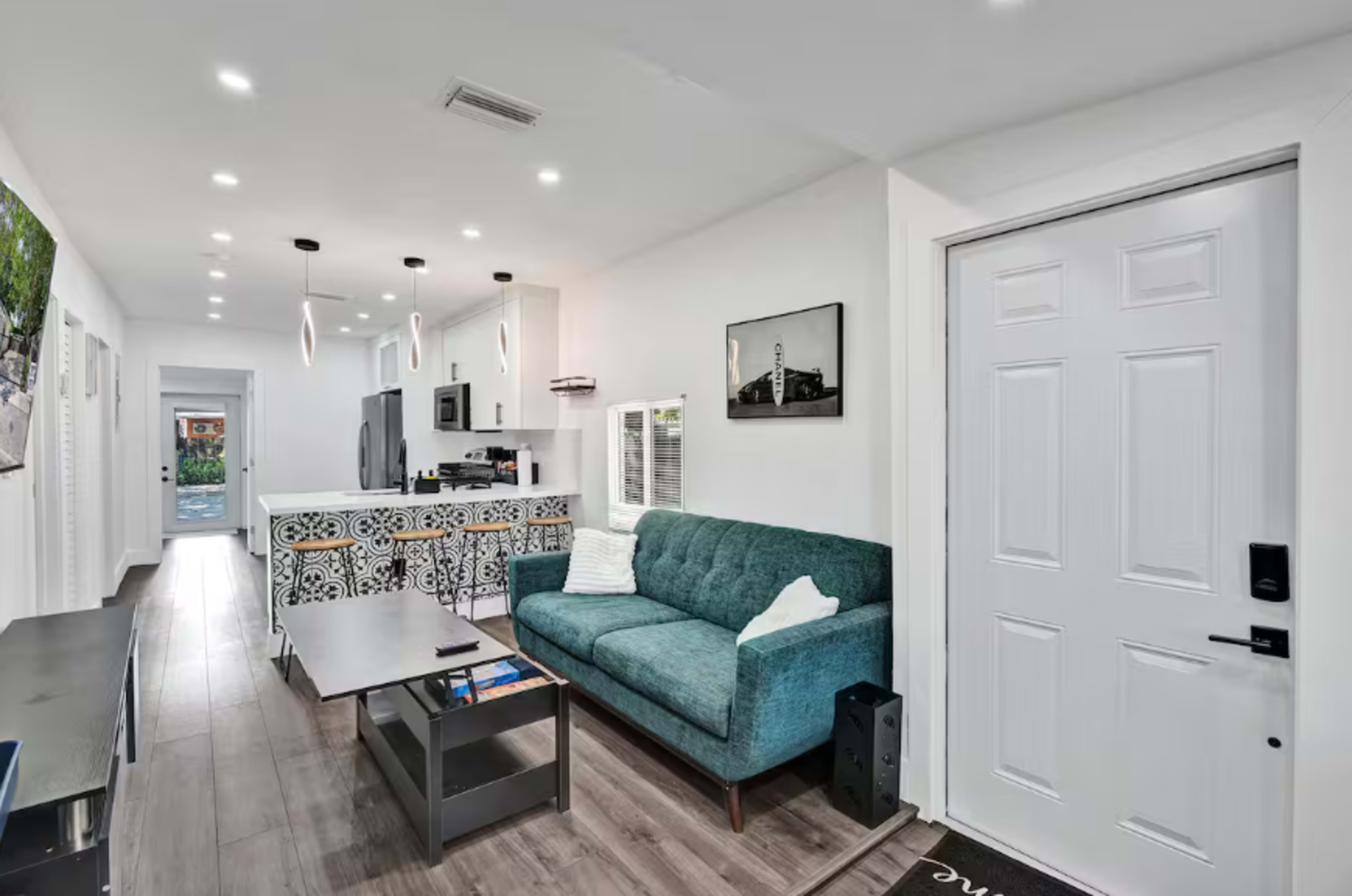 The image shows a modern living area featuring a teal couch, a coffee table, and a kitchen with a decorative backsplash.