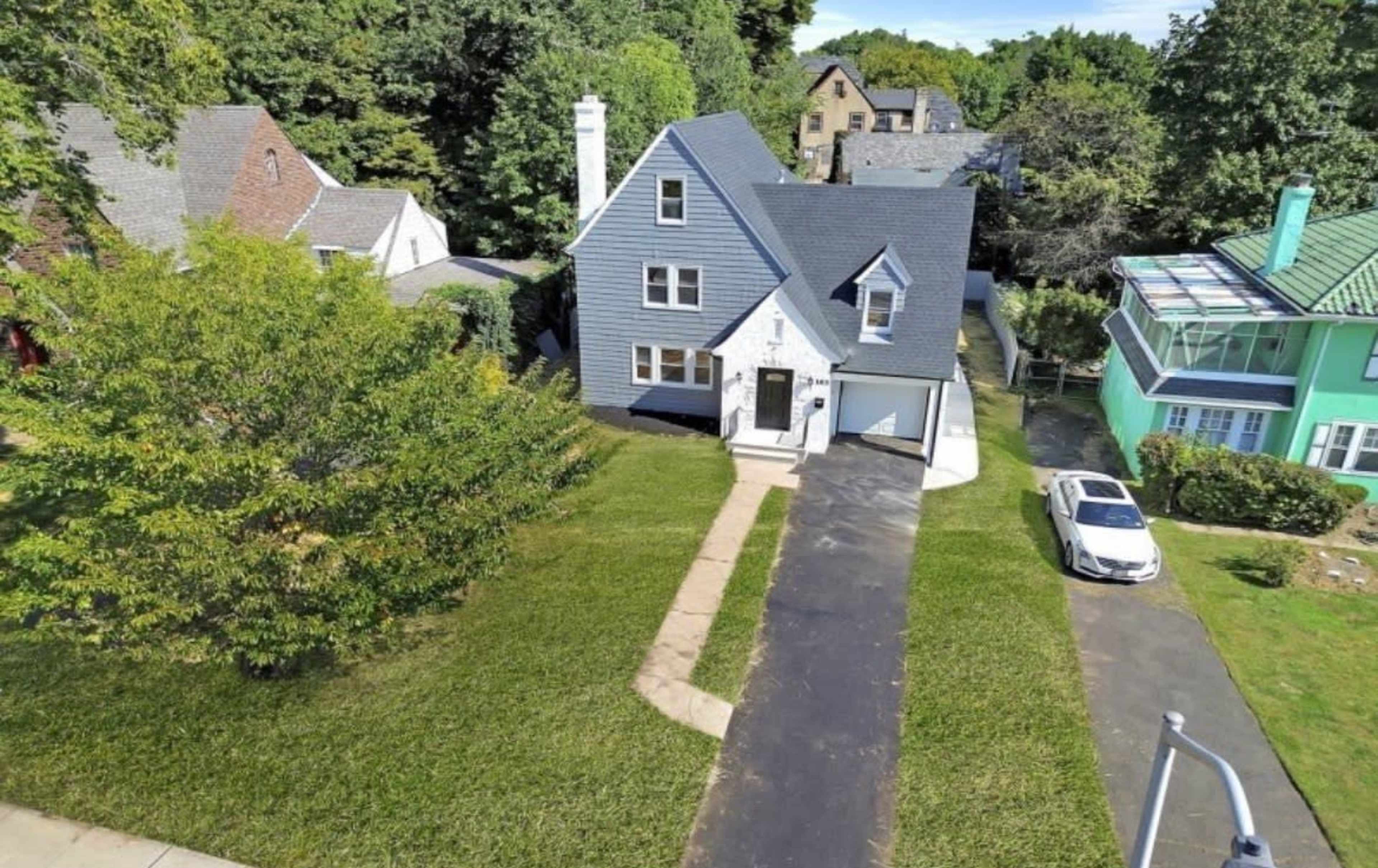 A two-story house with gray siding and a white door sits on a landscaped lot, surrounded by trees and other homes.