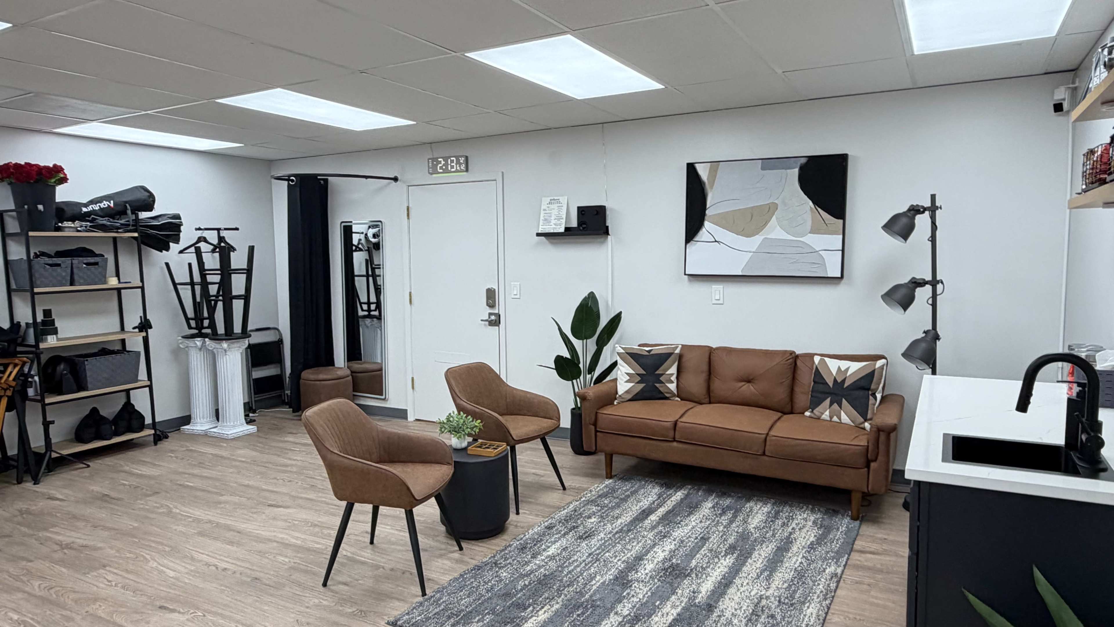 The image shows a modern, minimalist studio space featuring a brown couch, two chairs, a small table, and shelves with various items against a light-colored wall.