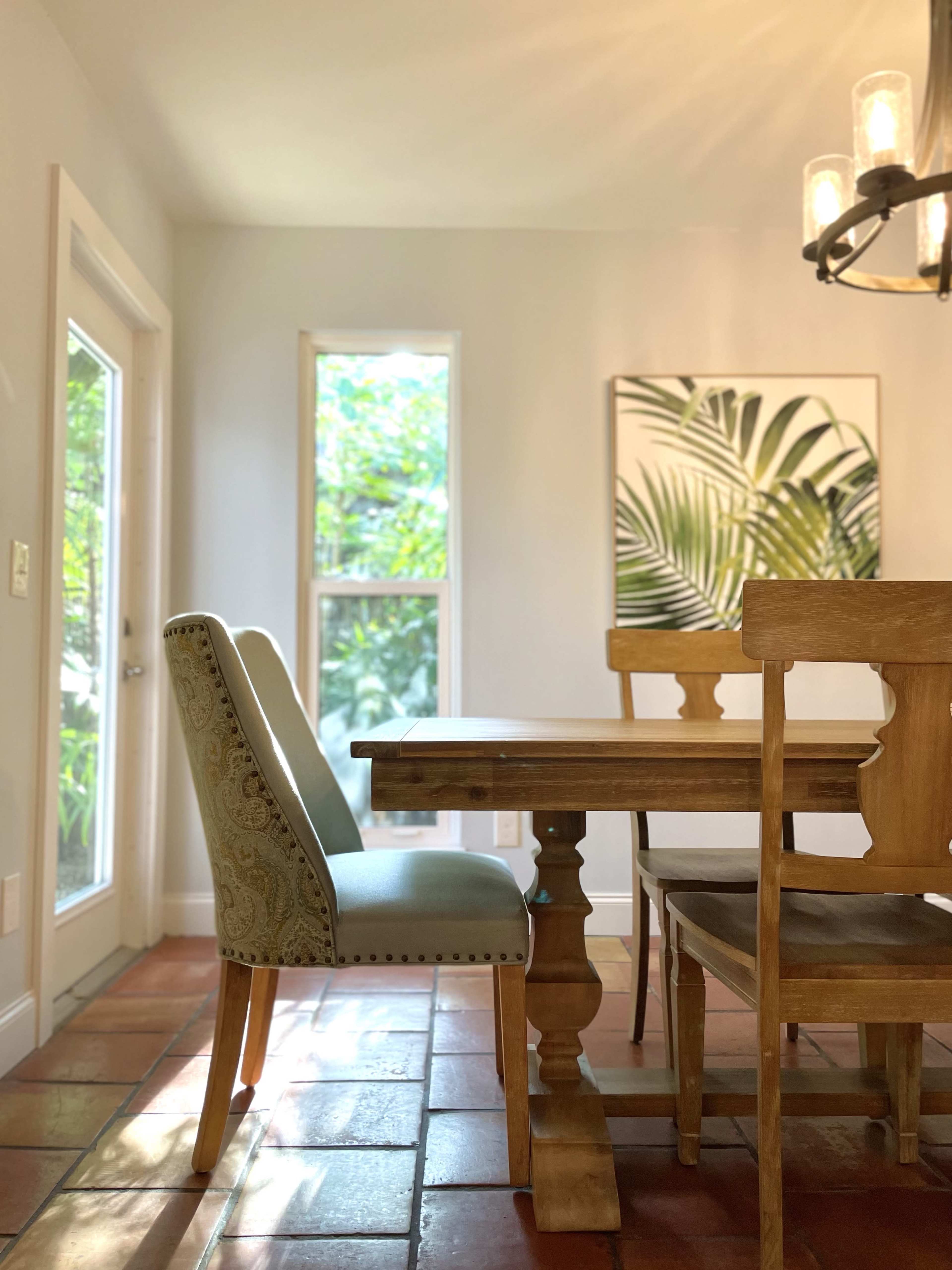 A wooden dining table surrounded by two chairs and a decorative chair, with a large window letting in natural light and a tropical-themed artwork on the wall.