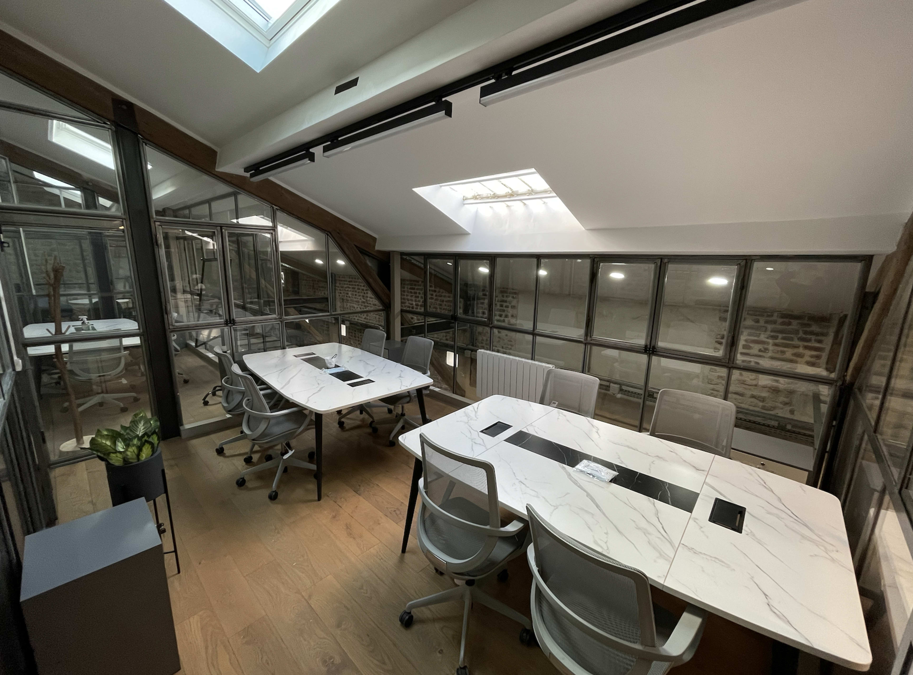 A modern office space with two white marble conference tables, surrounded by glass walls and wooden beams.