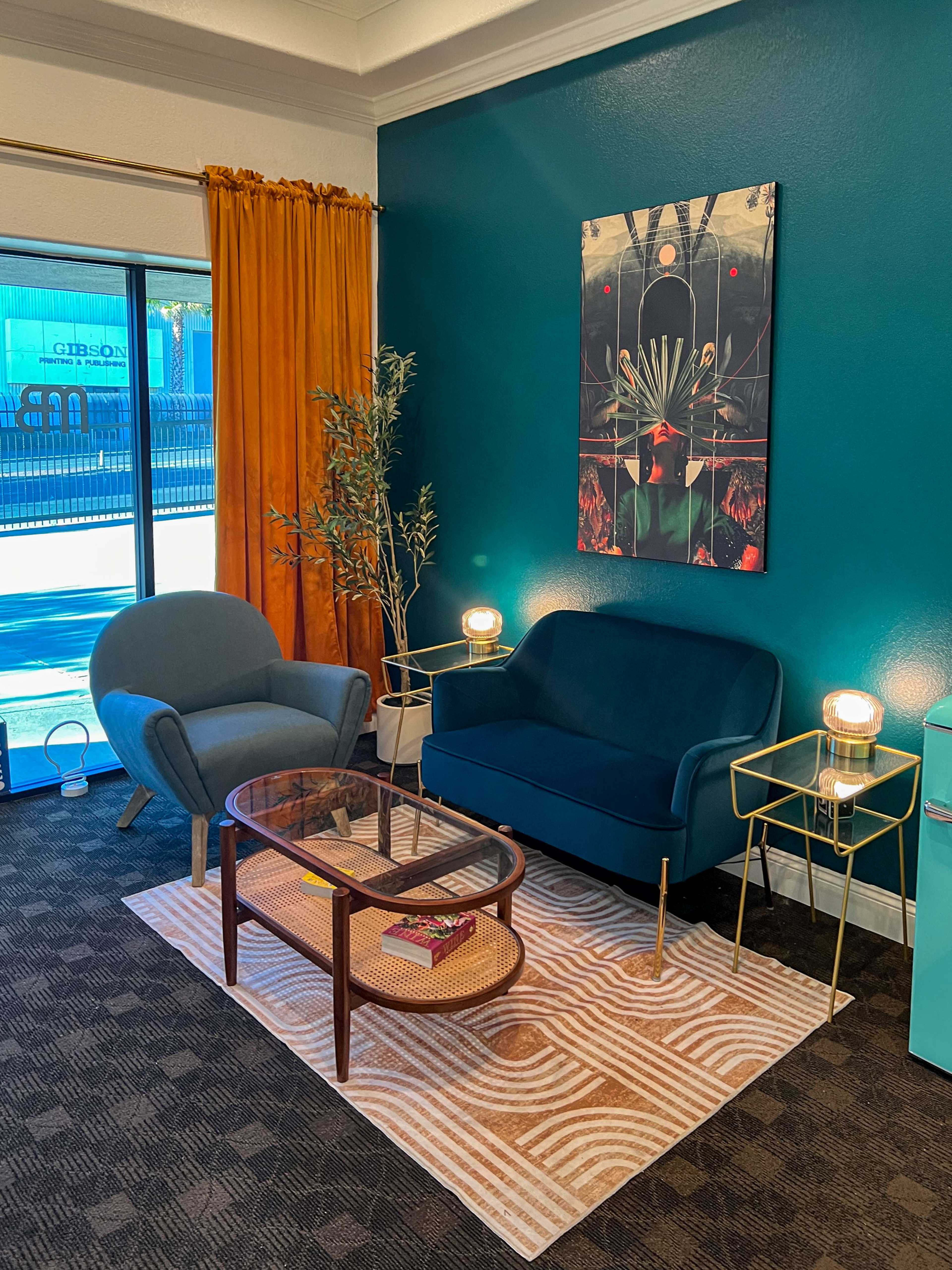The image shows a cozy seating area with a blue armchair, a green sofa, a glass coffee table, and two lamps, all set against a teal wall.