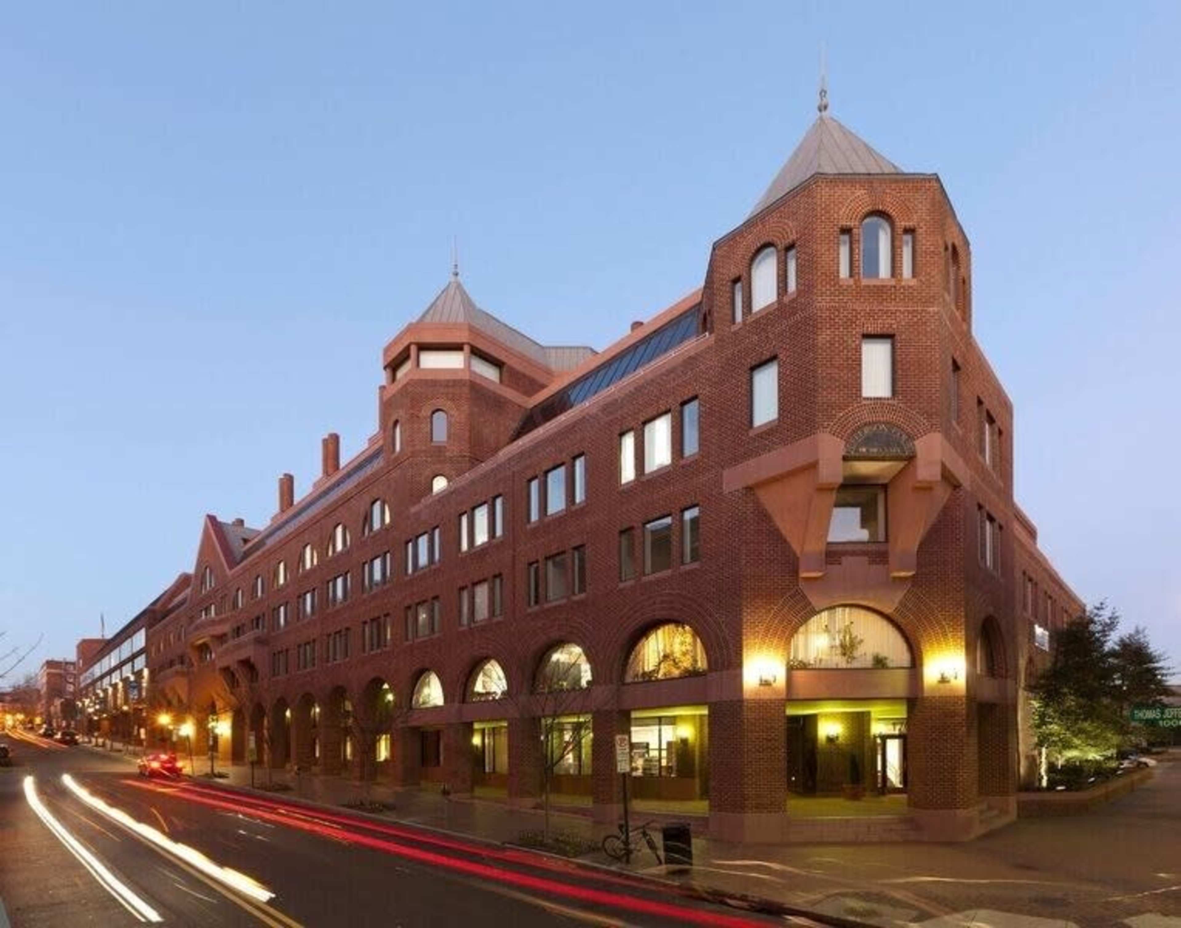 The image shows a large brick building with multiple arches and a peaked roof, situated on a street and illuminated by exterior lights during twilight.