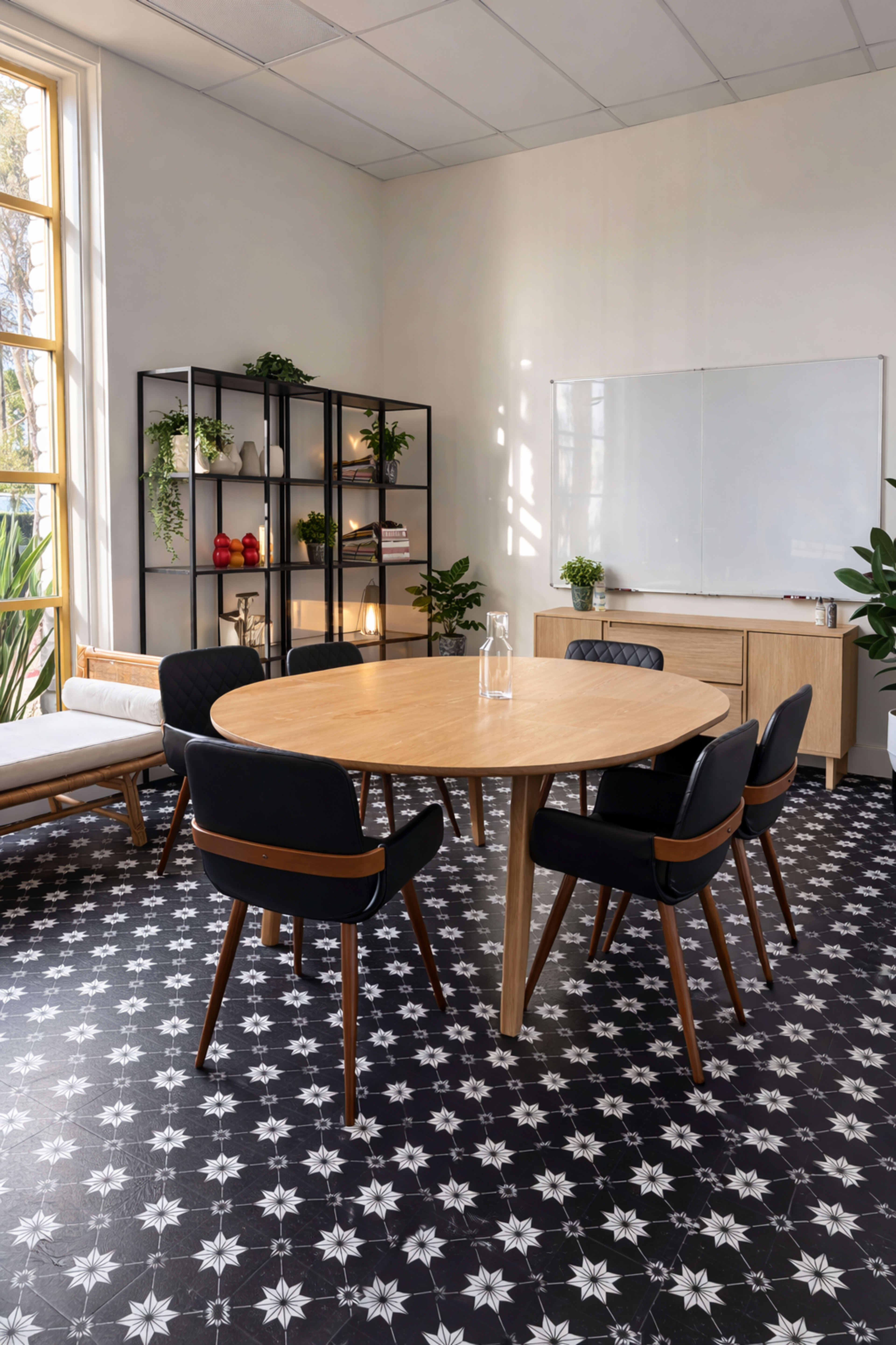 The image shows a modern conference room with a round wooden table surrounded by black chairs, a whiteboard on the wall, and a bookshelf filled with decorative items and plants.