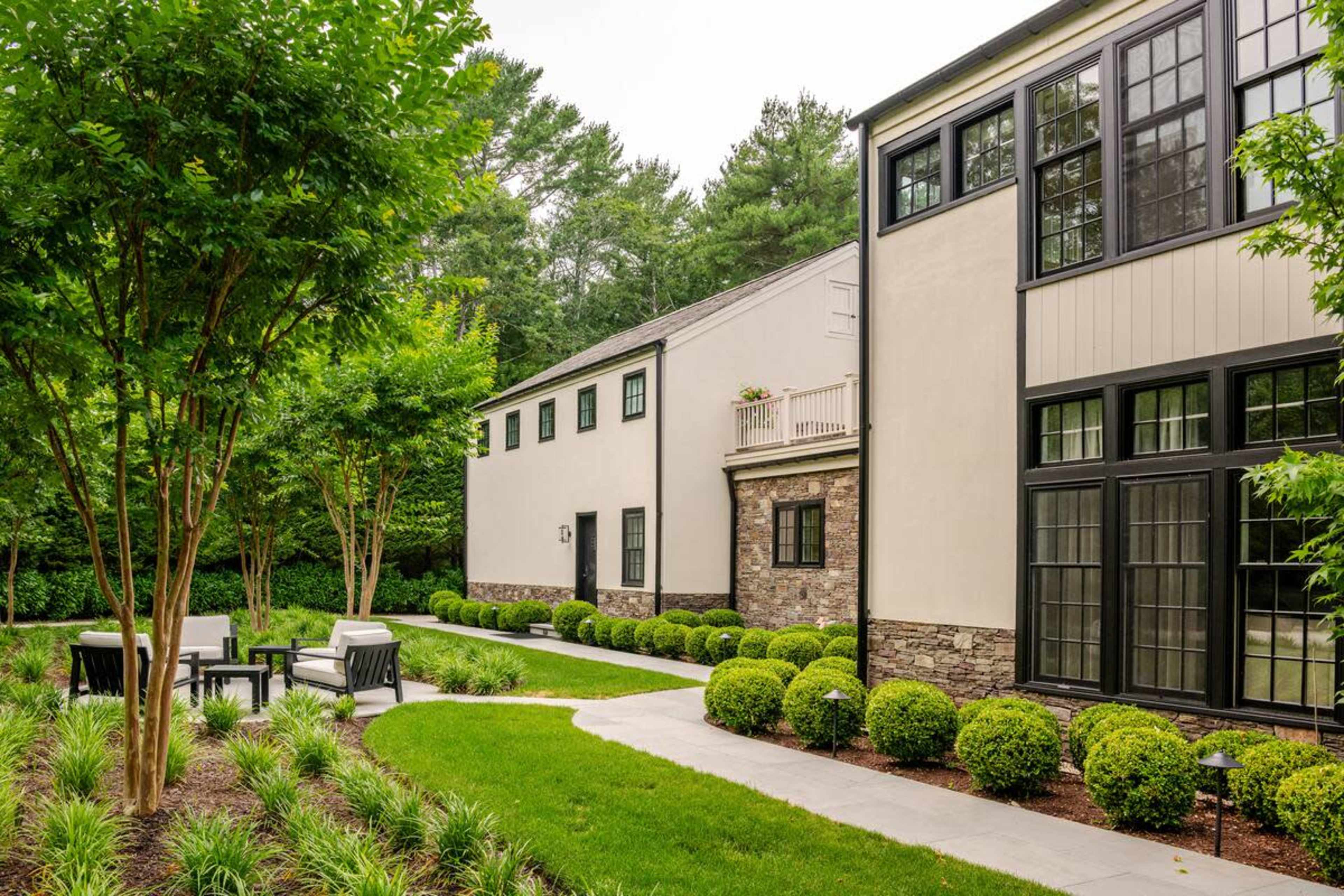 The image shows a well-manicured lawn with two modern buildings on either side and a stone pathway leading through neatly trimmed hedges and outdoor seating.