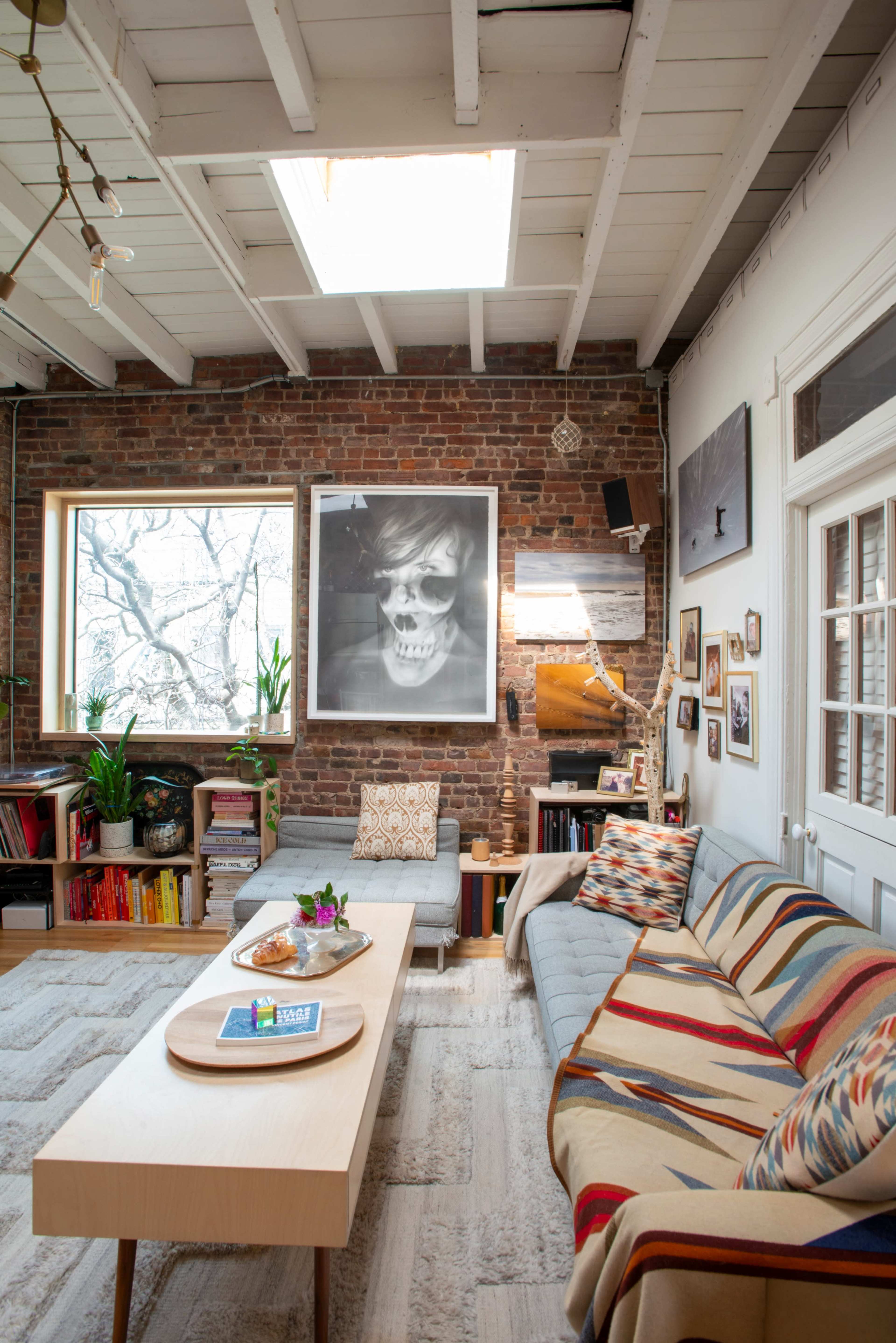 The image shows a modern living room with exposed brick walls, a large window, a gray sofa with a patterned blanket, a wooden coffee table, and artwork displayed on the walls.