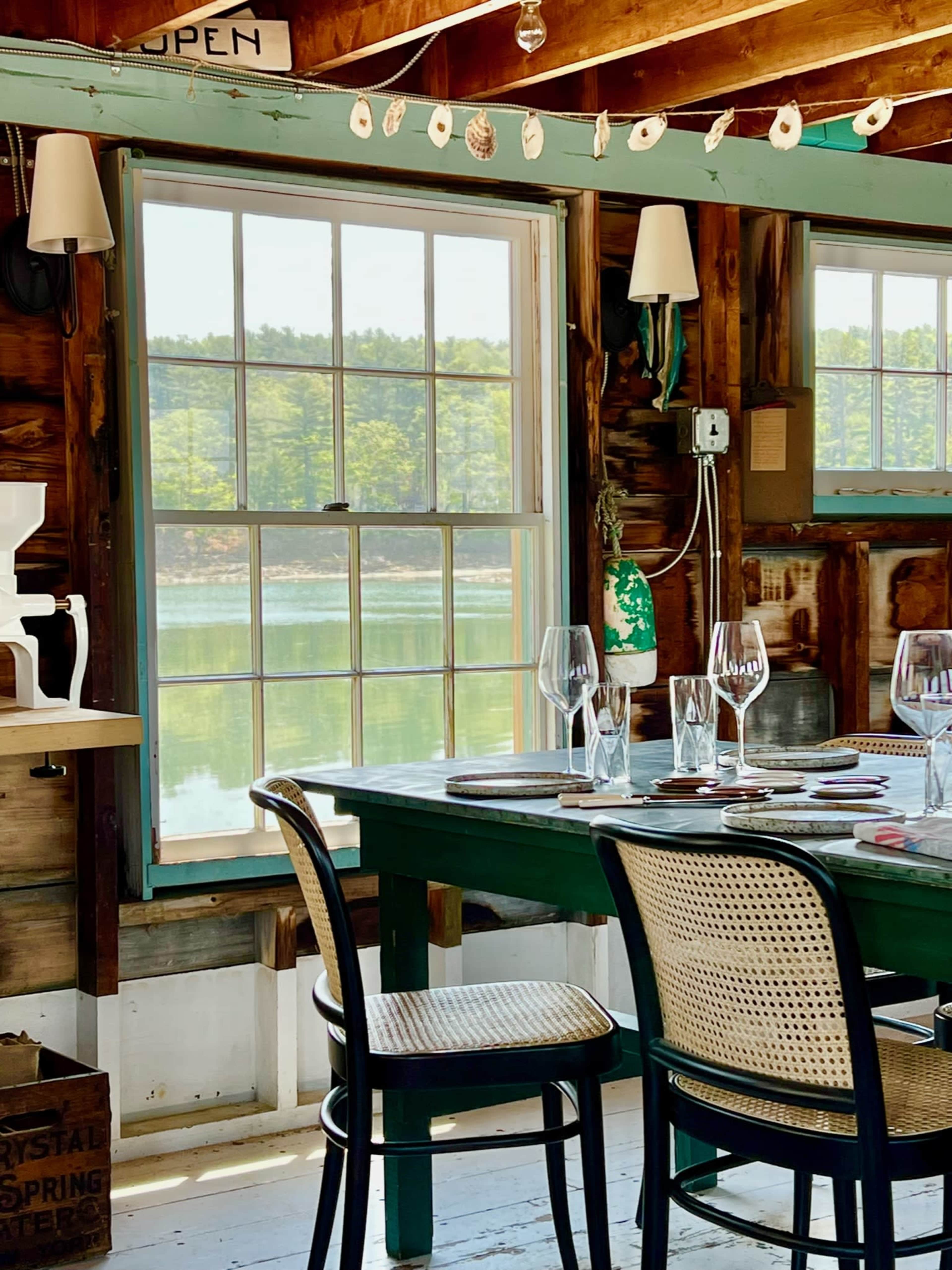 The image shows a rustic dining area with a green table set for a meal, featuring wine glasses, and a large window overlooking a body of water and trees.