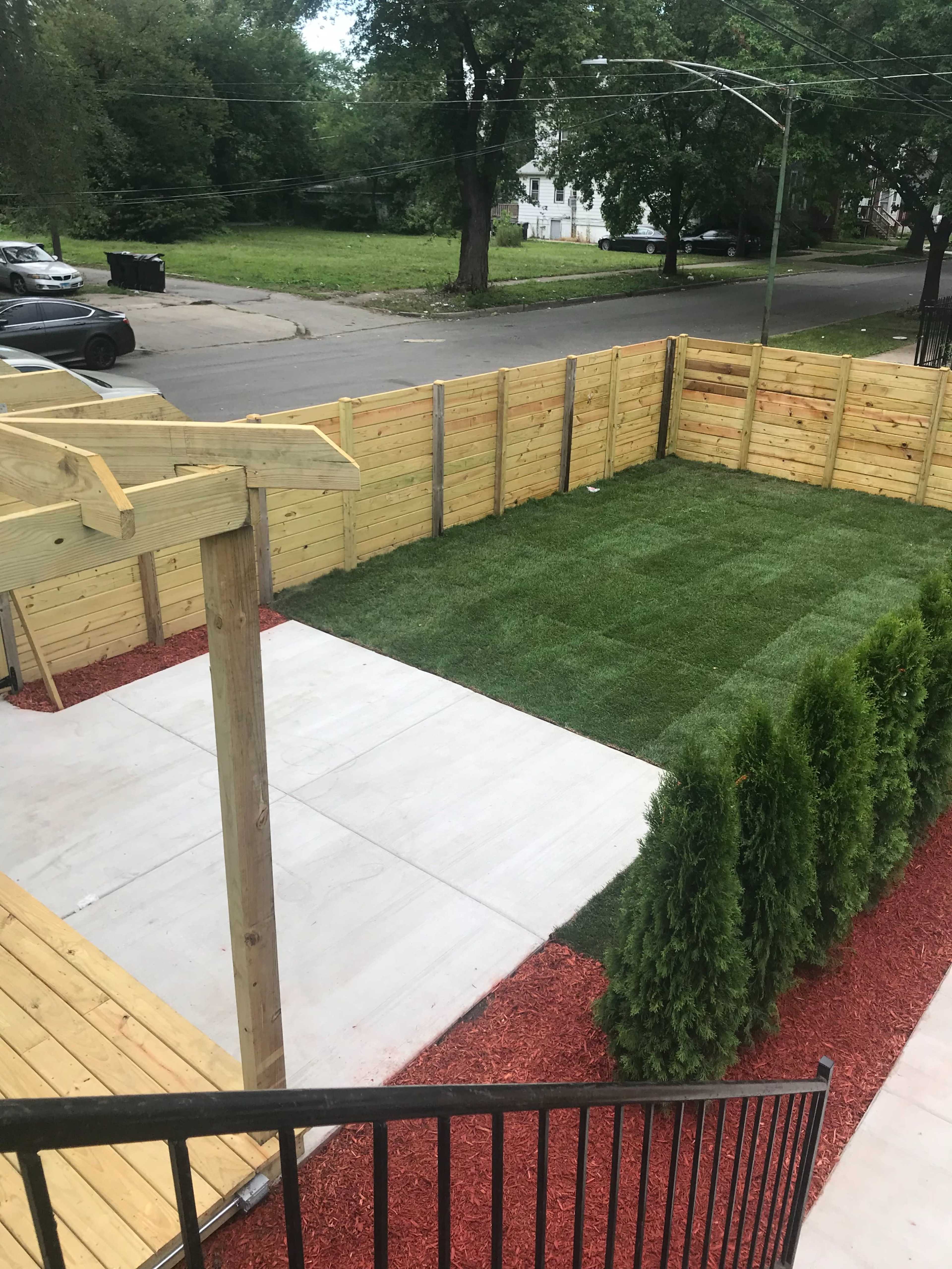 A fenced yard with a grassy area surrounded by wooden fencing and landscaping, featuring a concrete patio and red mulch.