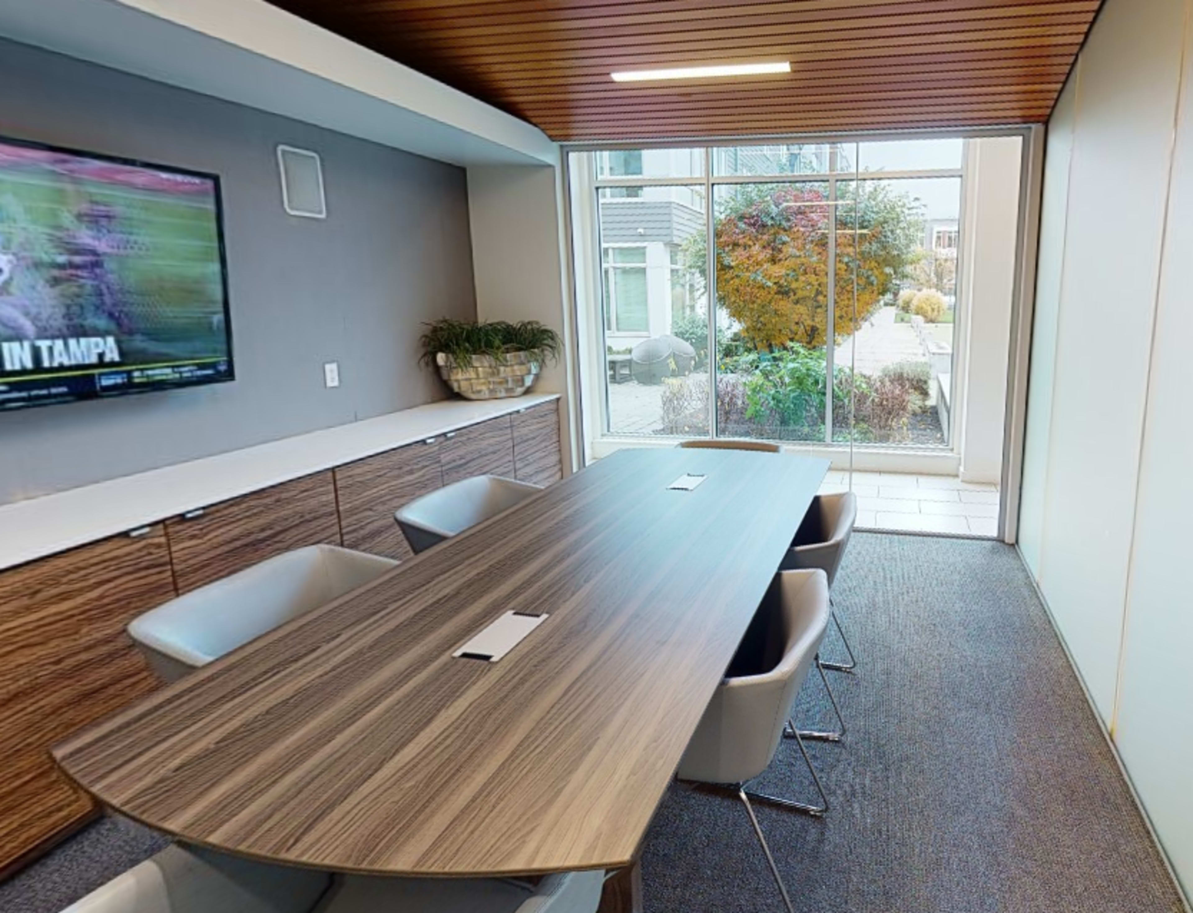 A modern conference room features a large wooden table, six white chairs, and a wall-mounted TV, with a view of a landscaped outdoor area through a large window.