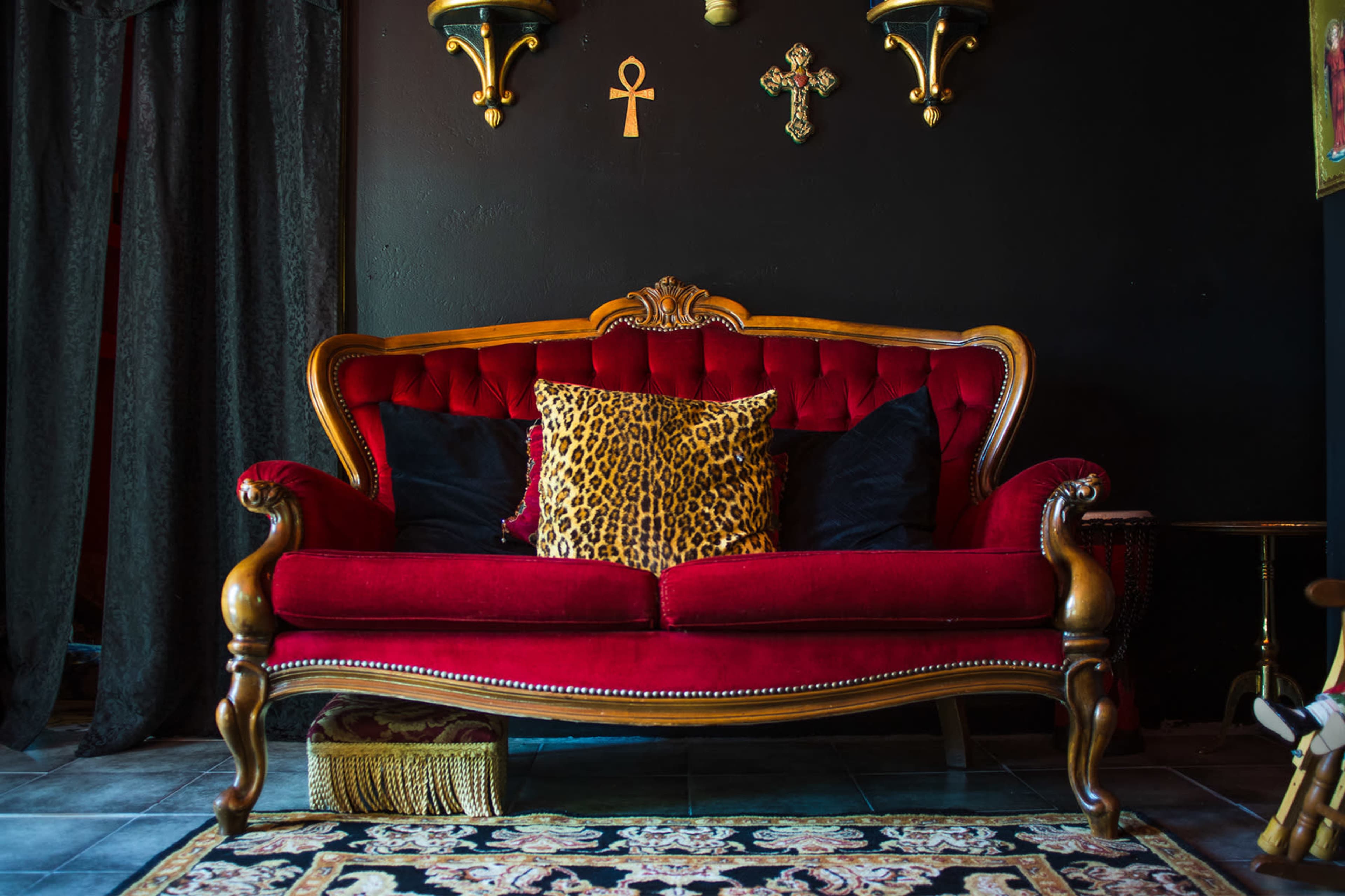 A red tufted sofa with wooden legs and ornate detailing is positioned in front of a black wall adorned with various wall decorations and framed art.