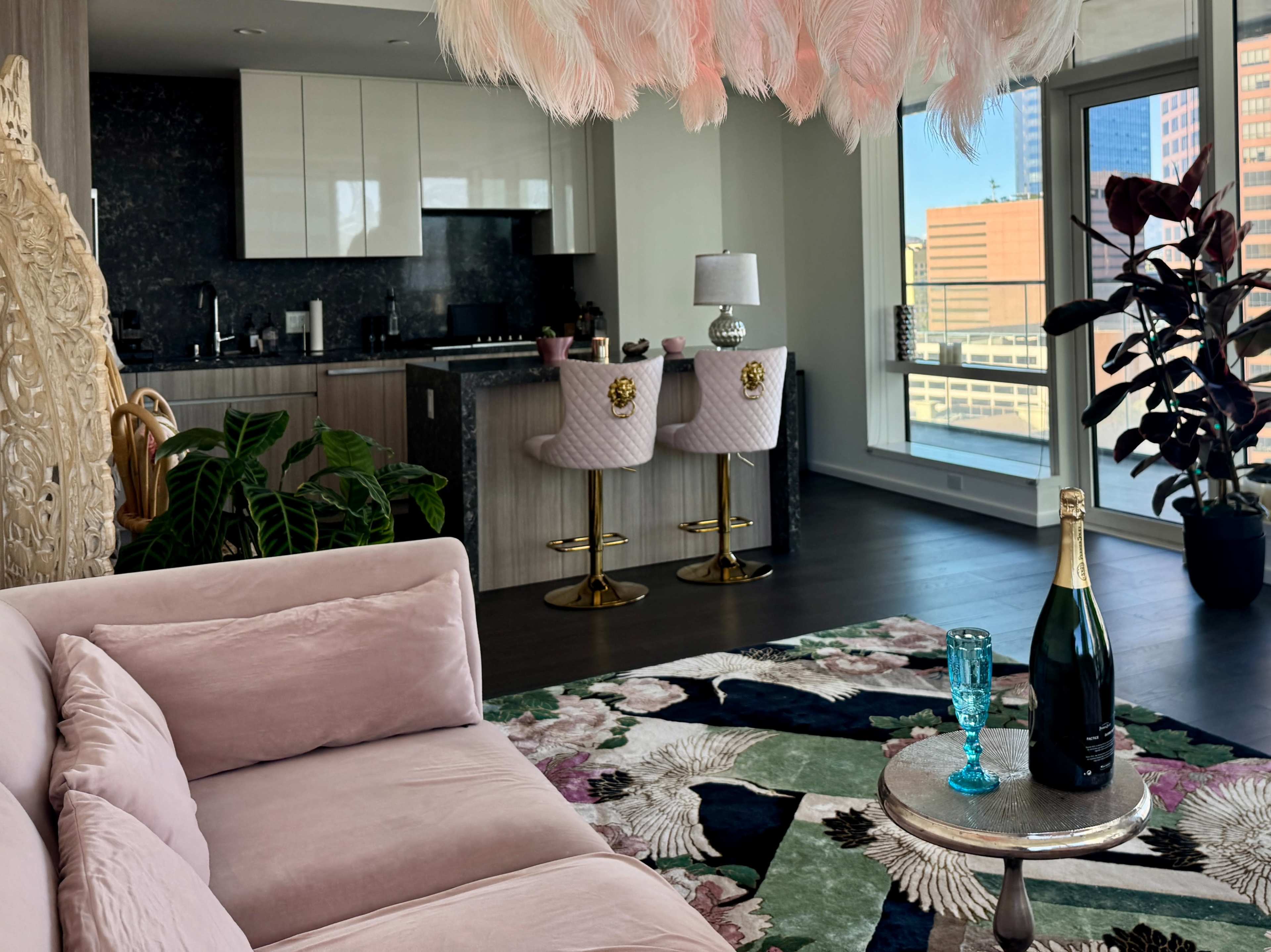 The image shows a modern living space featuring a pink sofa, a low table with a bottle of champagne, and a kitchen area with sleek cabinetry, alongside large windows that offer a city view.