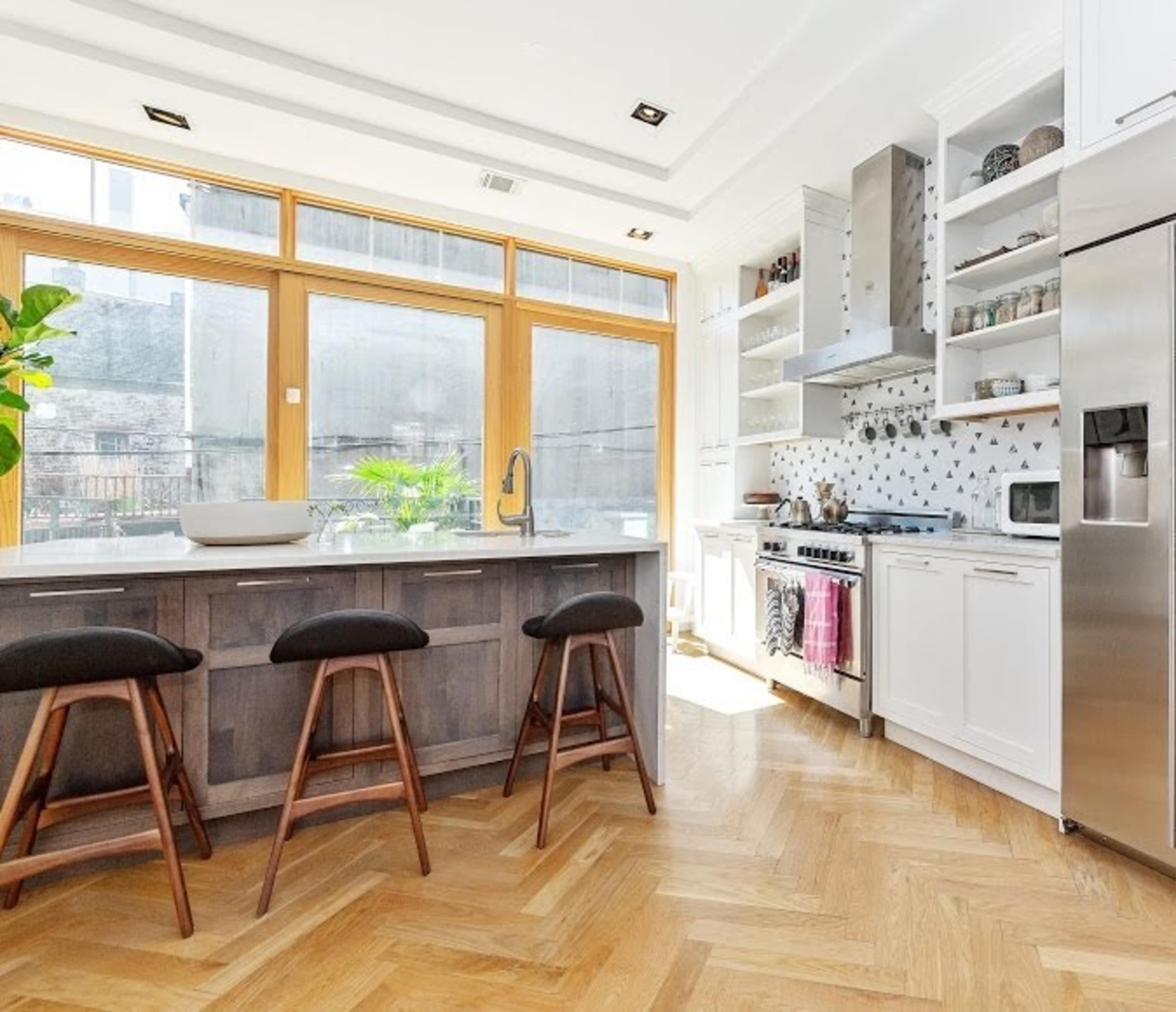 The image shows a kitchen with large windows, a central island with three stools, and modern appliances against a wall decorated with patterned tiles.