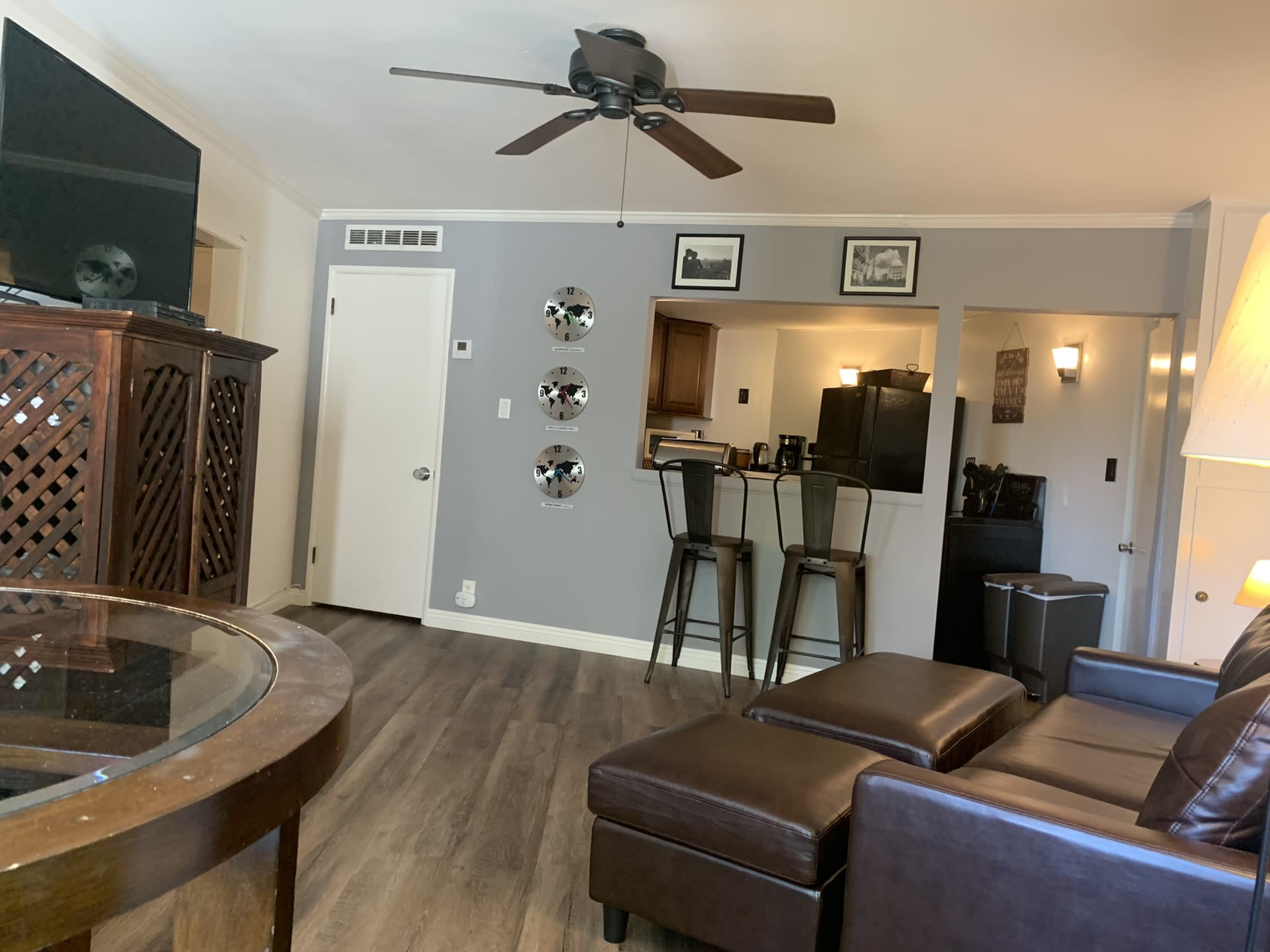 A modern living room with a dark leather sofa, a wooden coffee table, a TV mounted on the wall, and a kitchen area with bar stools visible in the background.