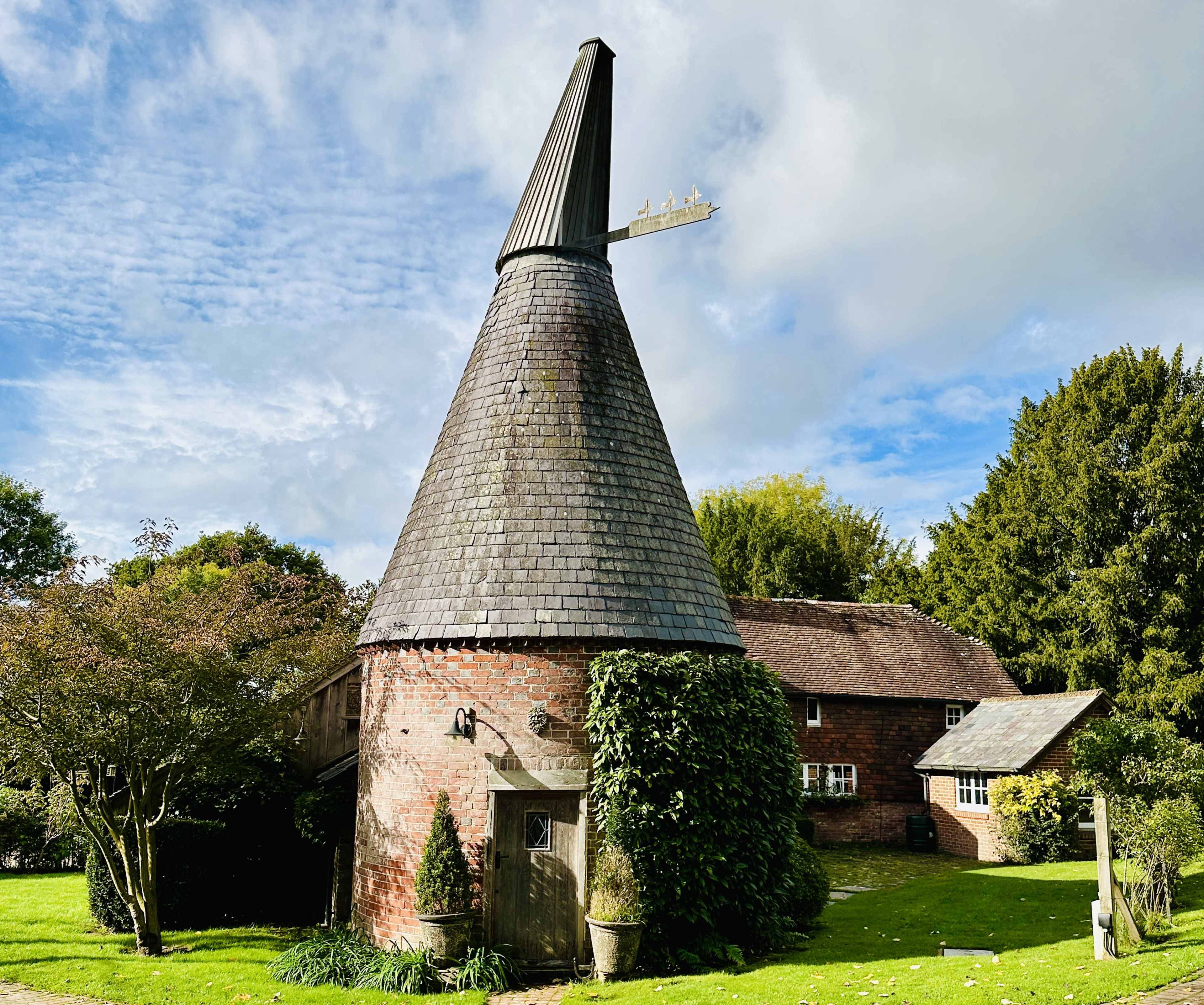 A traditional oast house with a conical roof is surrounded by greenery and set against a cloudy sky.