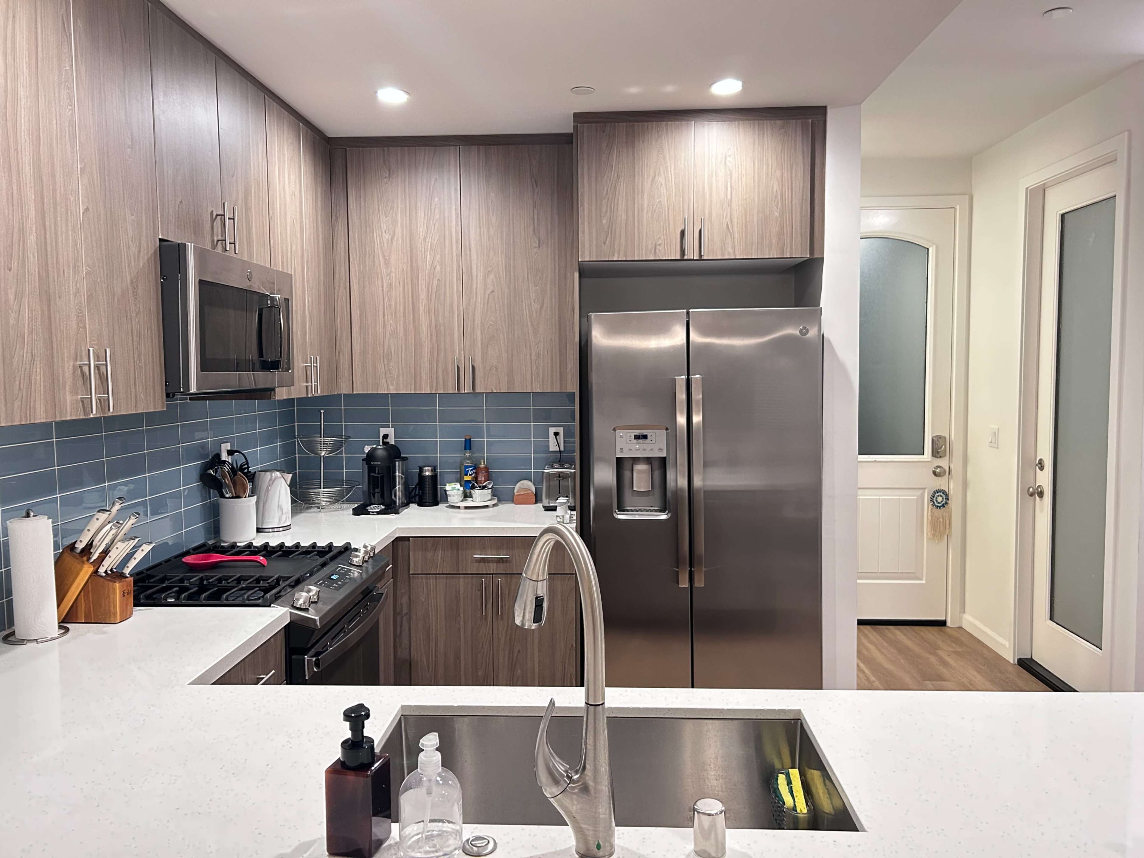 The kitchen features modern cabinets, stainless steel appliances, and a blue tile backsplash, with a view of an entrance door.