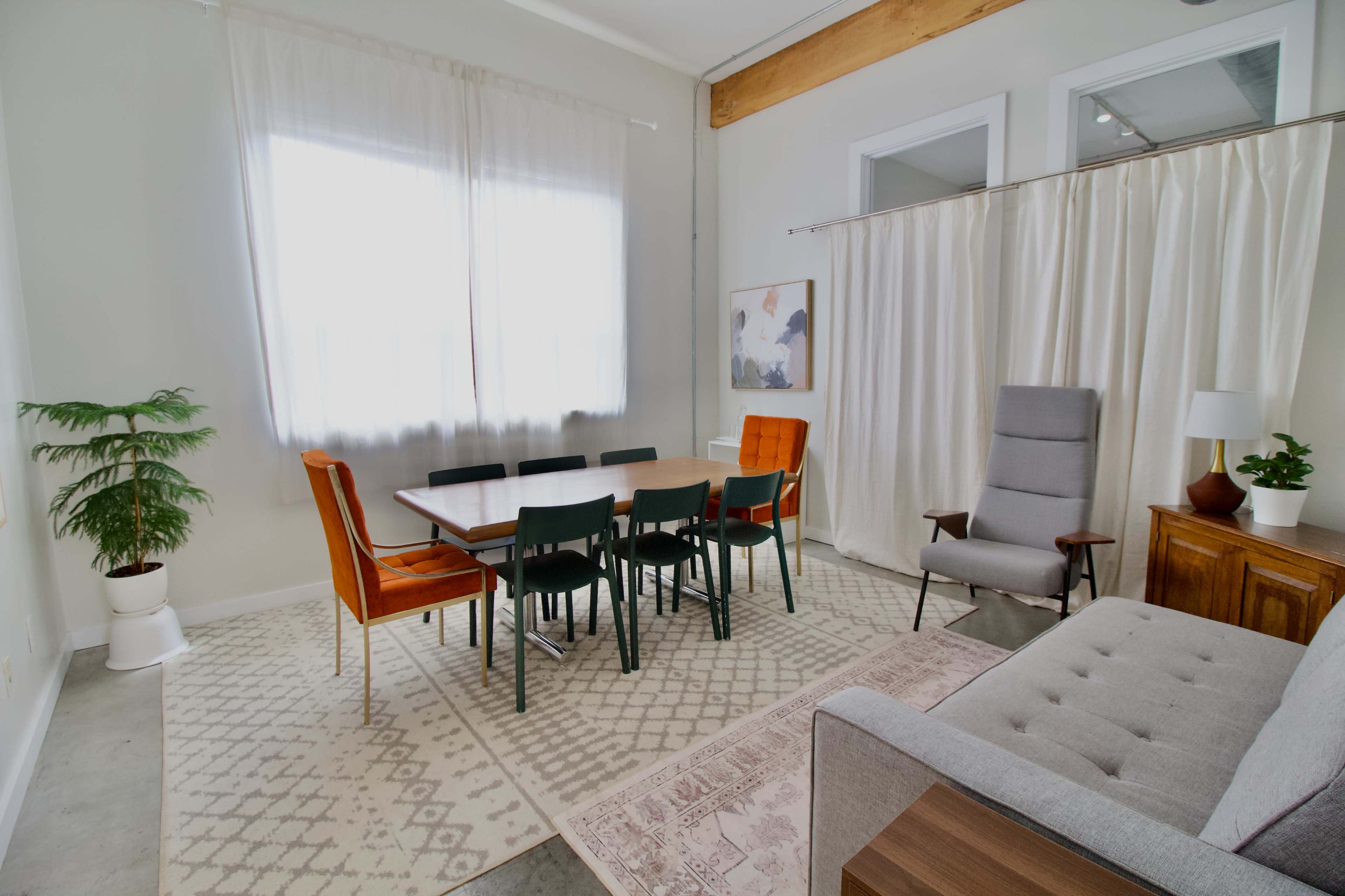 The image shows a bright, modern dining area with a large table surrounded by chairs, a gray sofa, and a potted plant.