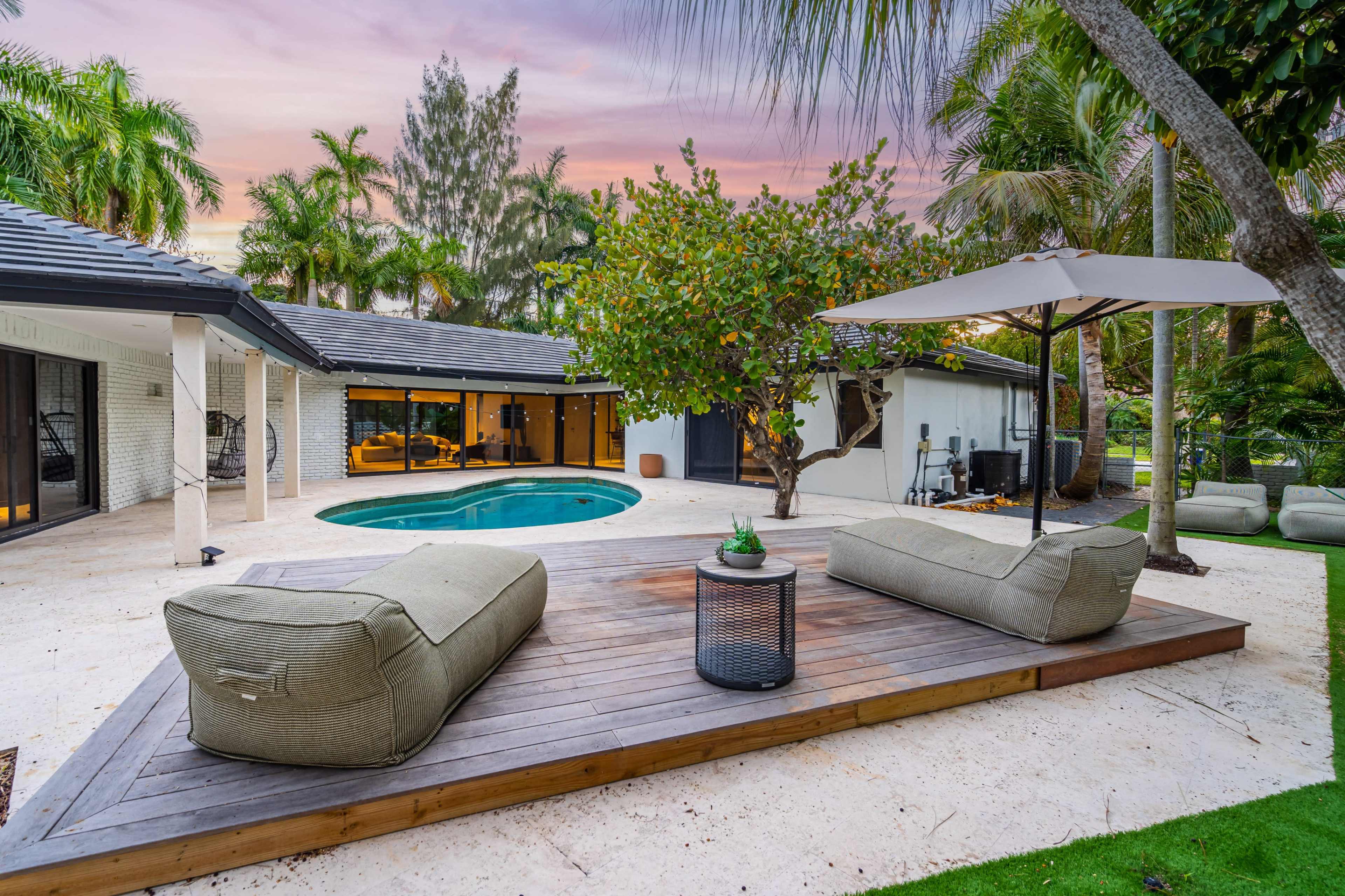 The image shows a tranquil outdoor space featuring a swimming pool, lounge seating on a wooden deck, and lush tropical landscaping.