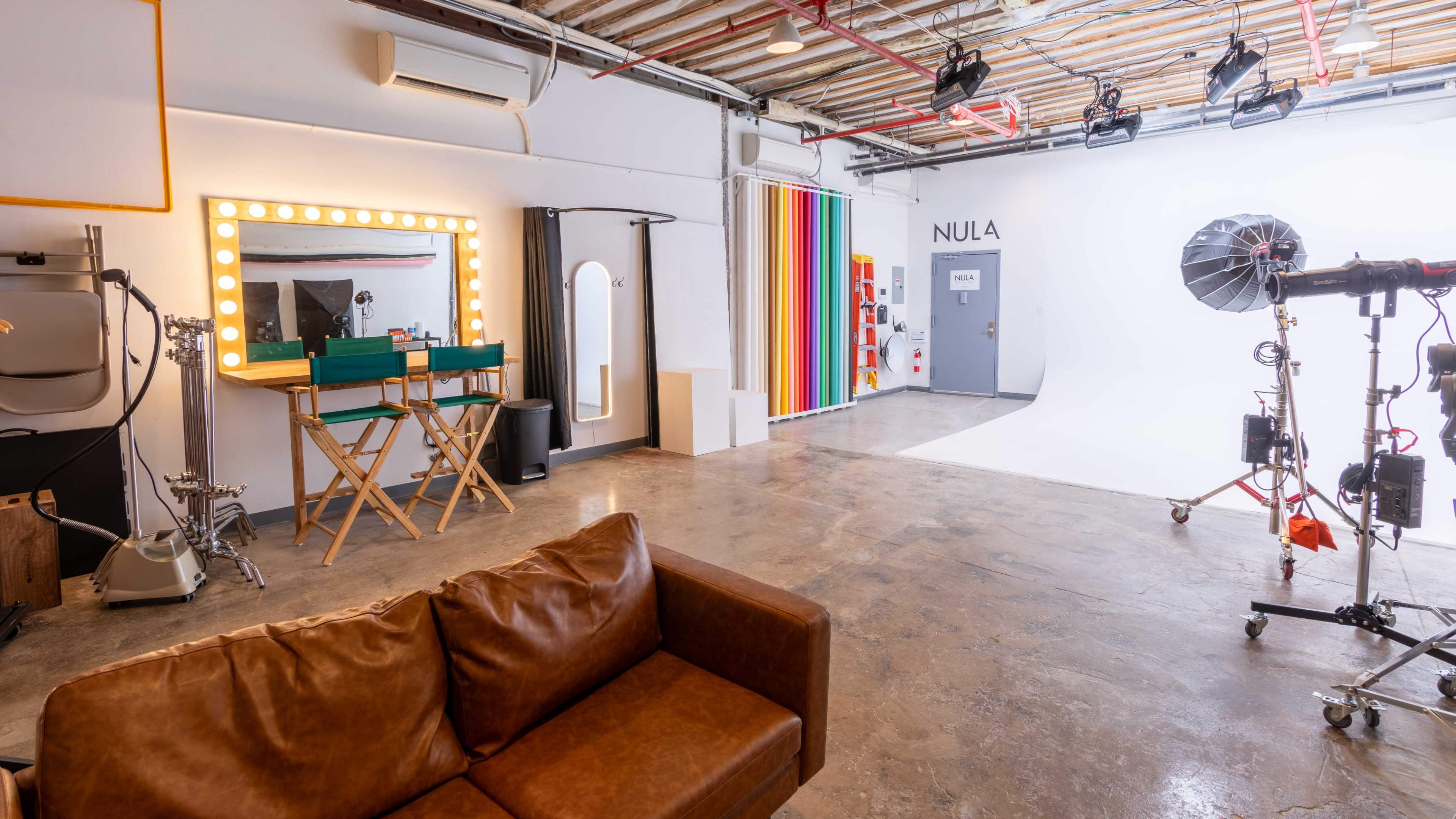 A bright studio space featuring a makeup mirror with lights, two chairs, a colorful striped wall, and various photographic equipment.