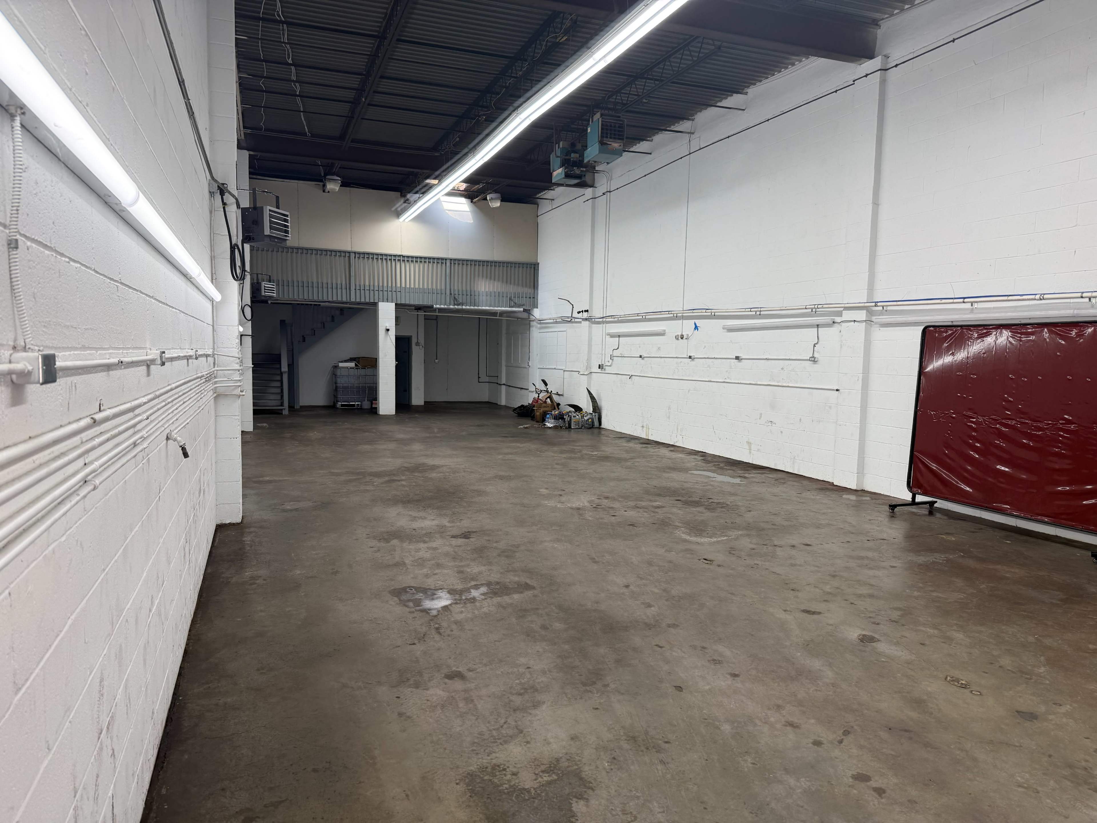 The image shows a large, empty industrial space with concrete flooring, white-painted walls, and overhead lighting.