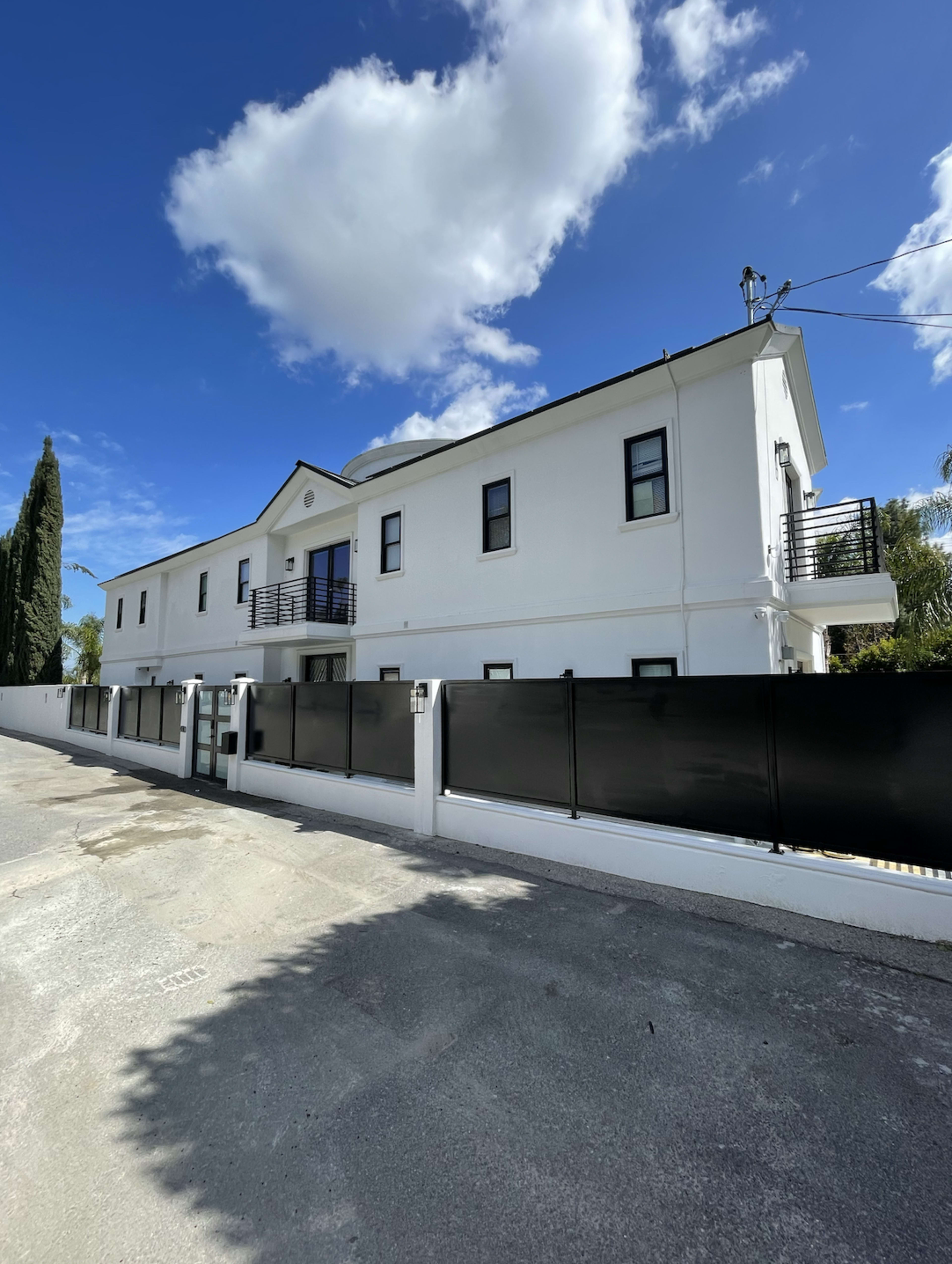 A modern two-story white house with black railings and a fence, set against a blue sky with clouds.