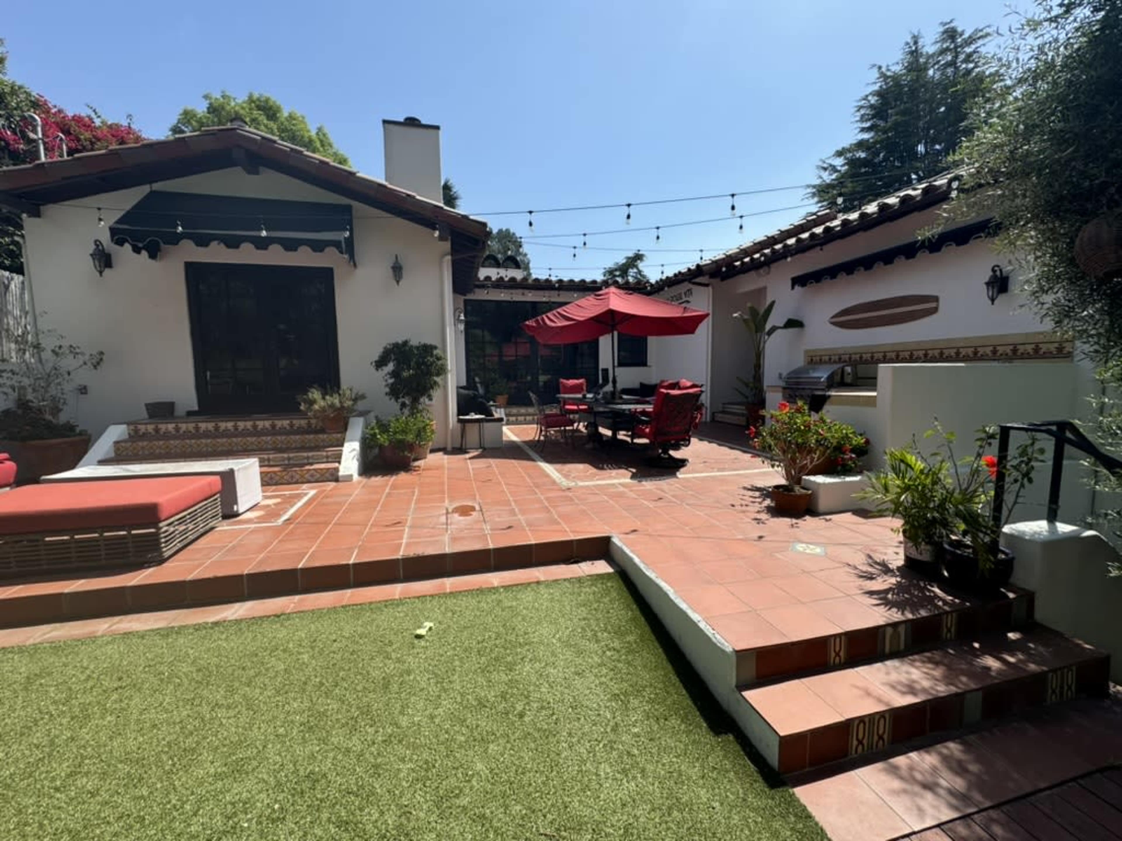A patio area features terracotta tiles, outdoor seating under a red umbrella, and greenery in pots, with adjacent white-walled buildings.