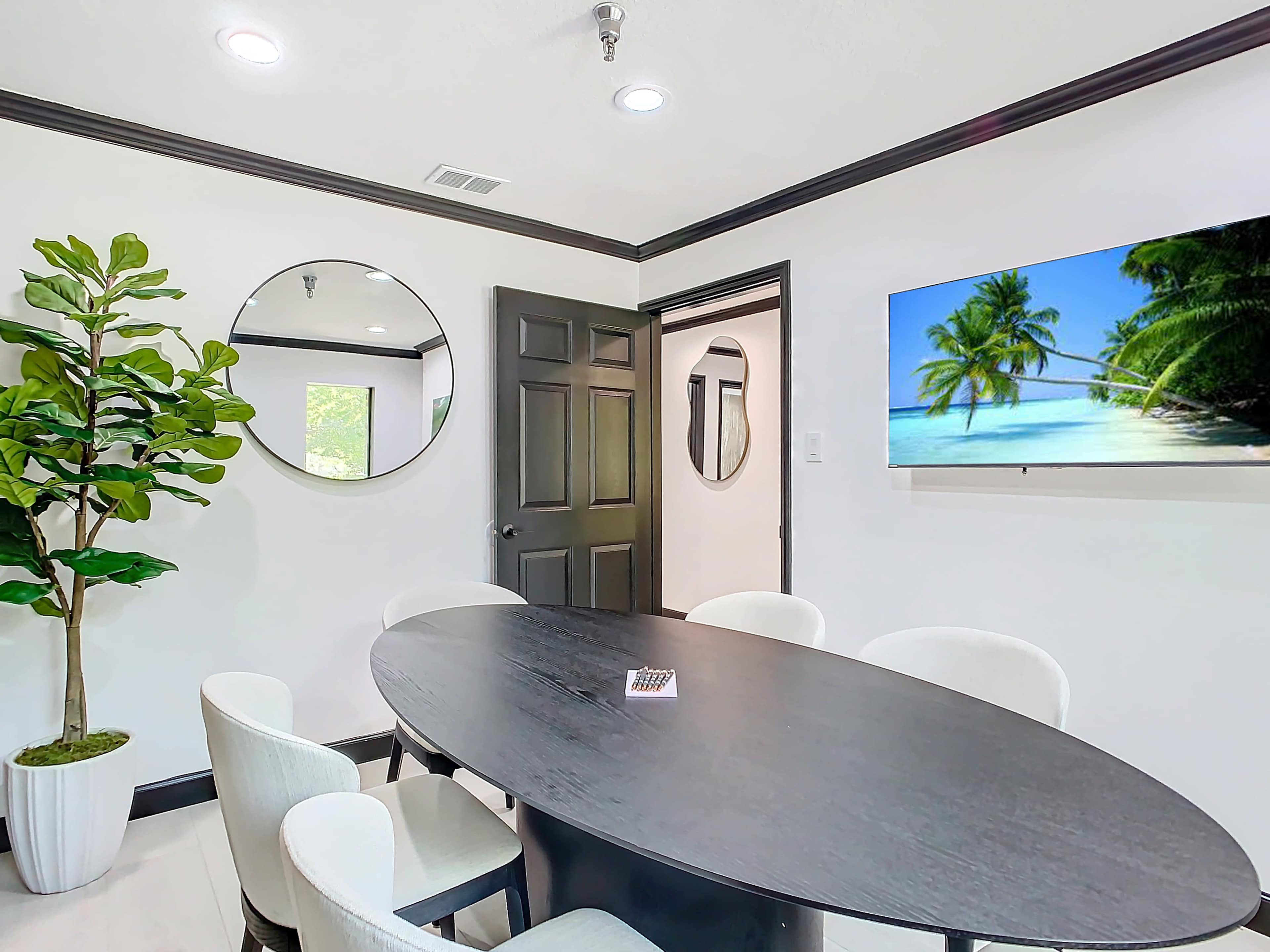 A round black dining table surrounded by white chairs is positioned in a bright room featuring a large mirror and a beach-themed TV display.