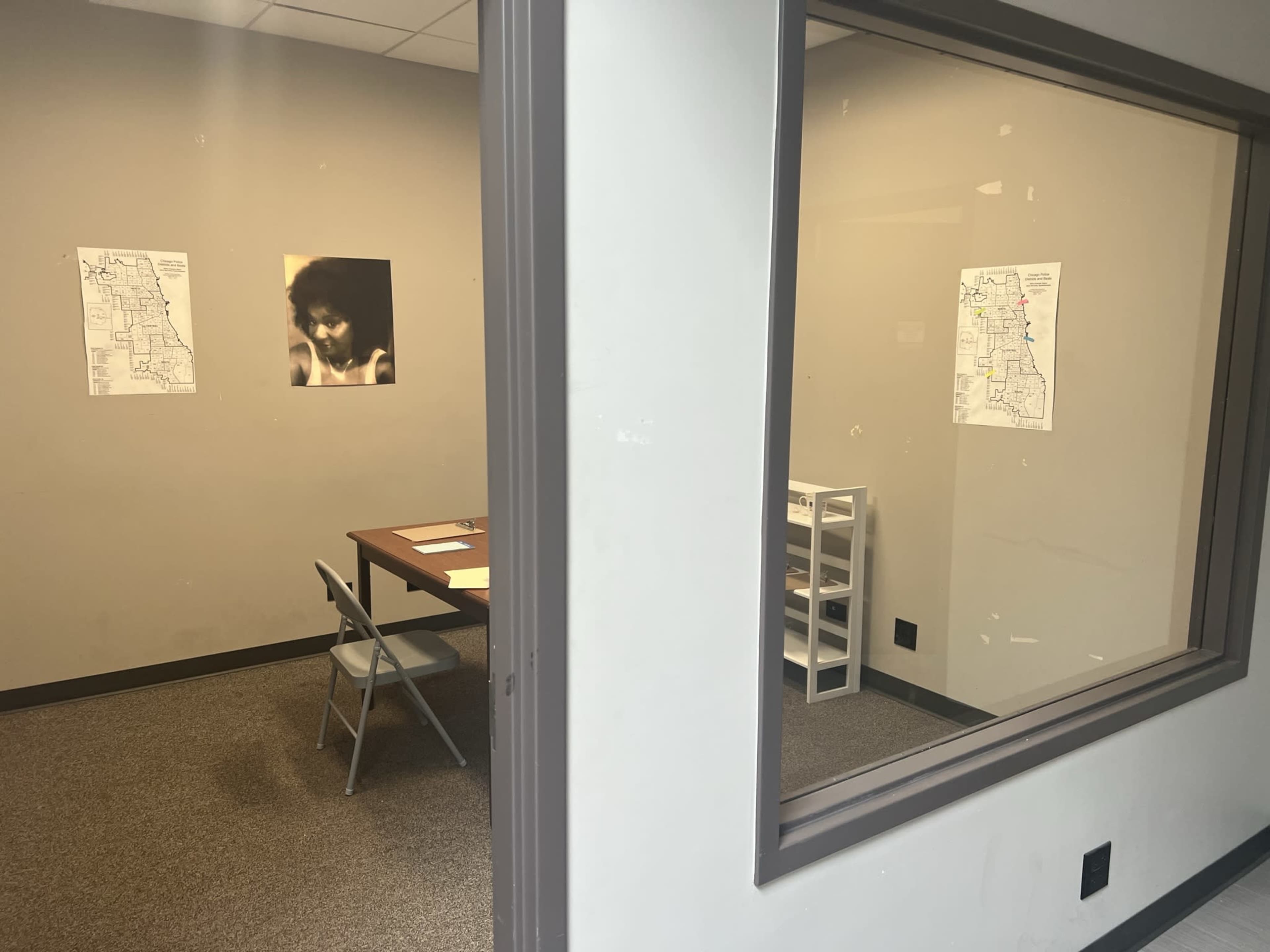 A table with chairs is set up in a room with two maps on the wall, visible through a large window from the adjacent space.