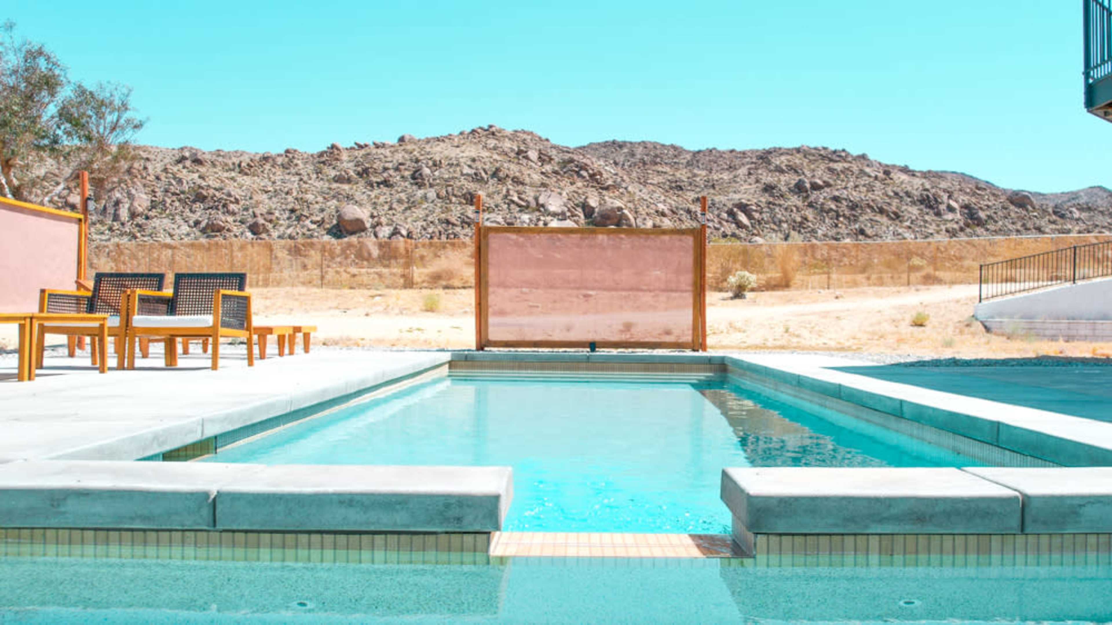 The image shows a swimming pool surrounded by stone tiles and wooden chairs, set against a backdrop of dry, rocky terrain and clear blue skies.