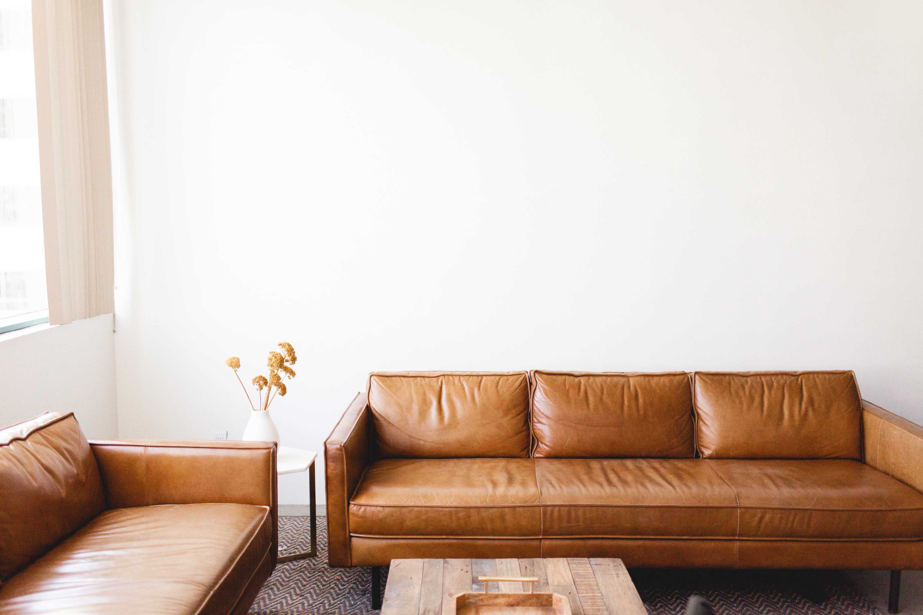 A light-filled room features two brown leather sofas and a wooden coffee table, along with a vase containing dried flowers.