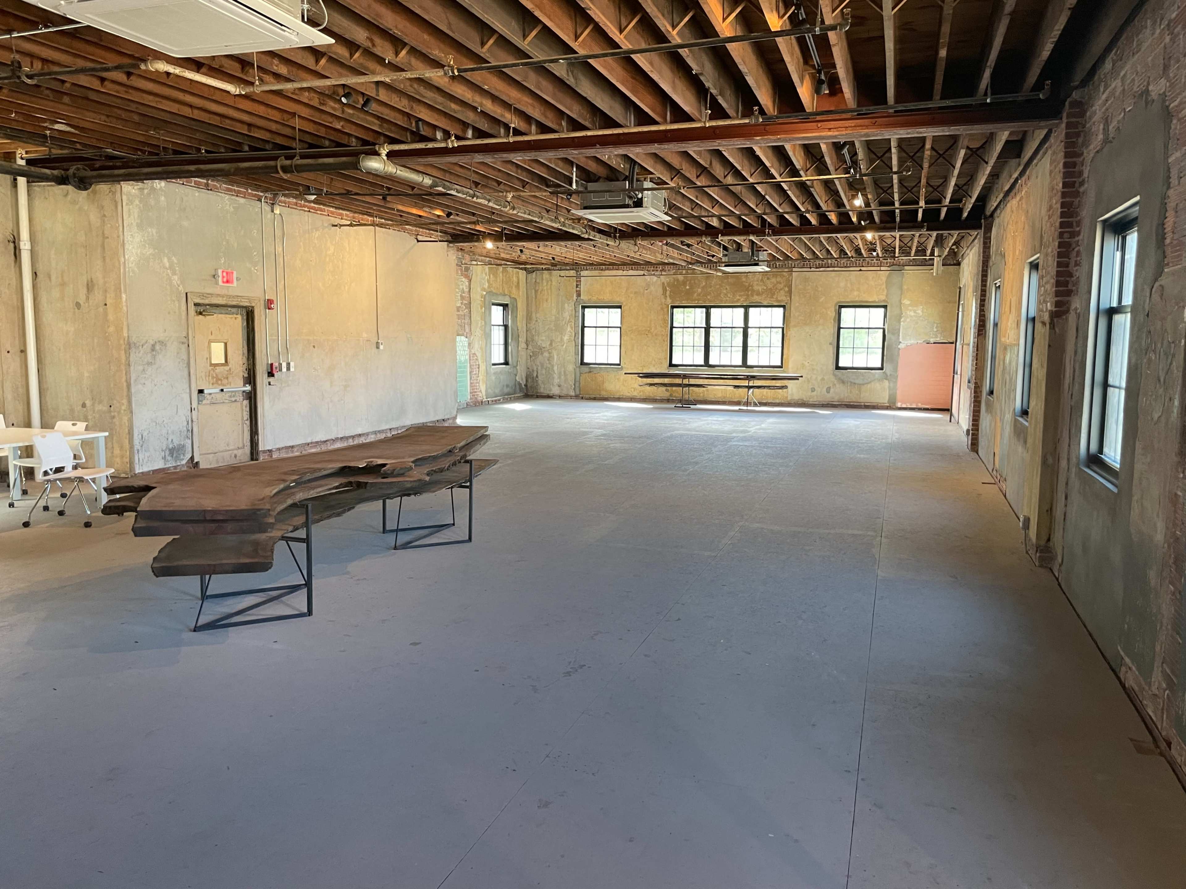 An empty room with exposed wooden beams, concrete flooring, and windows allowing natural light, featuring a large wooden table in the center.